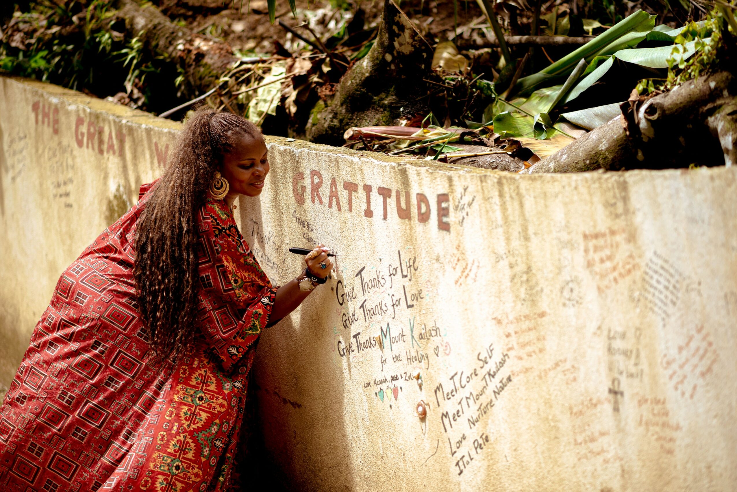 Woman with long curly hair writing on a wall with trees and plants above, wall has words like 'gratitude' and messages.