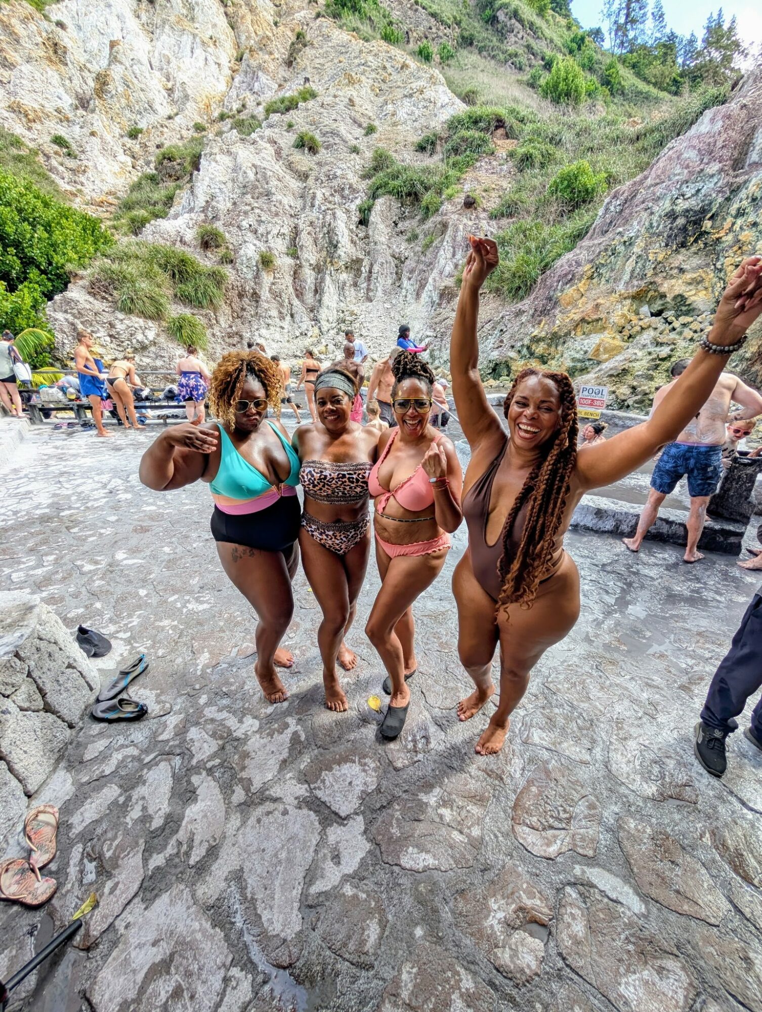 Four women in swimsuits standing on a rocky riverbank, smiling and celebrating, with cliffs and other people in the background.
