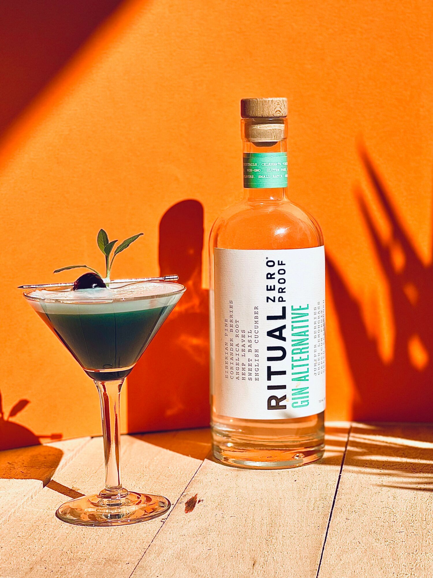 A bottle of Ritual Zero Alcohol Gin and a cocktail glass with a dark drink and a small green garnish, set against an orange background.