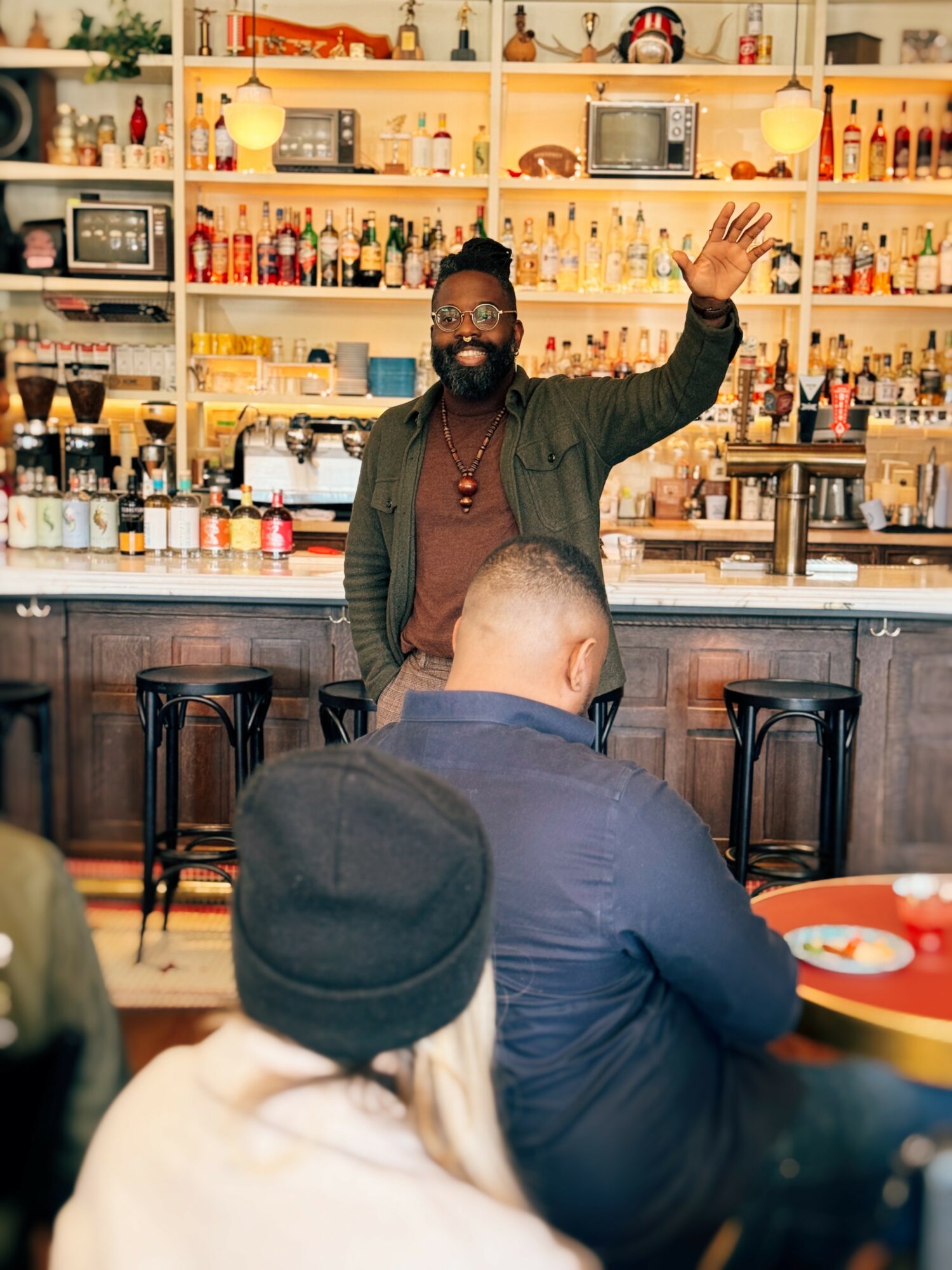 Man with glasses and beard waving in a bar with shelves of bottles behind him, people seated at tables in foreground.