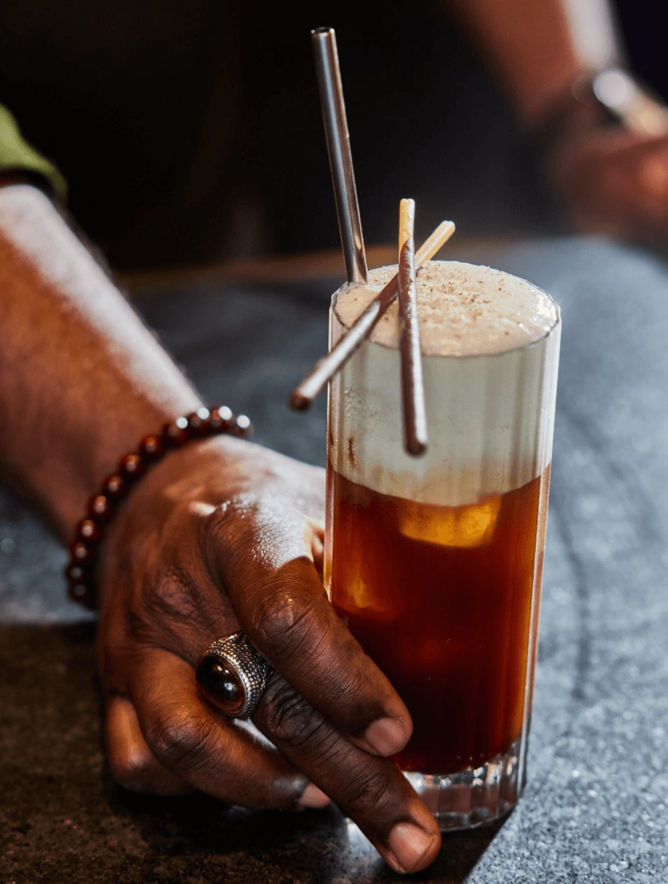 Person's hand with a ring and bracelet holding a tall glass of layered coffee drink with foam and straws.