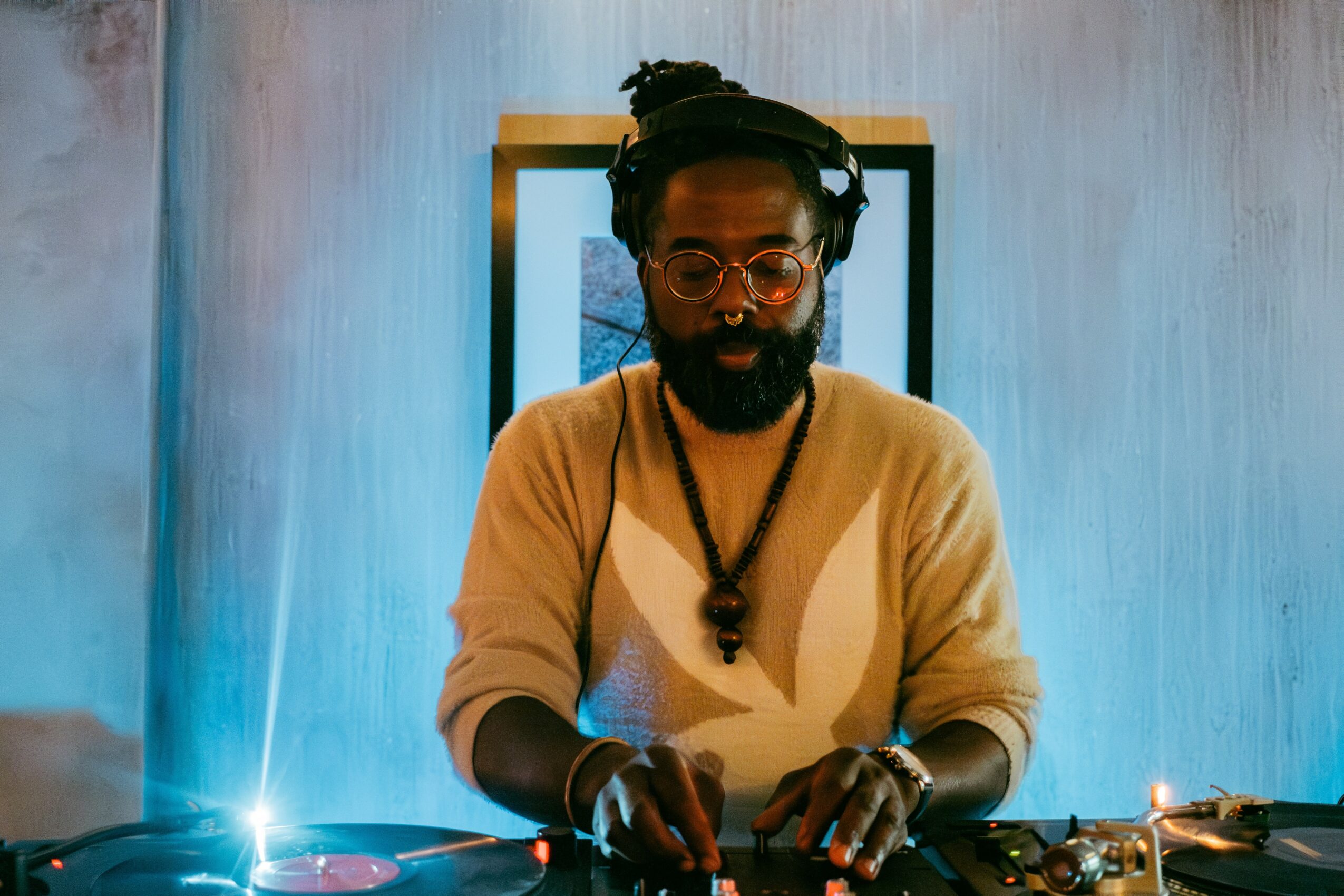 Person with glasses and dreadlocks wearing a beige sweater, DJing with turntables, in a room with blue lighting.