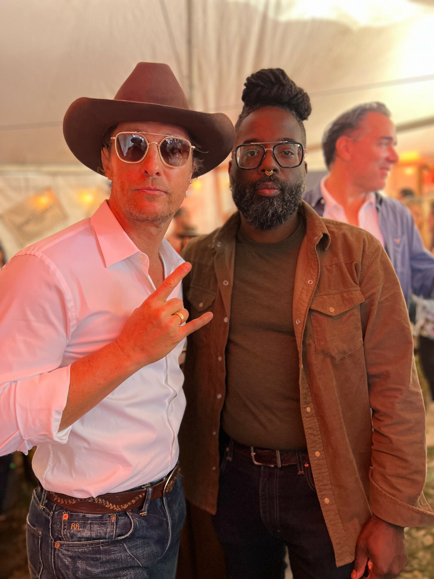 Two men standing together indoors, one wearing a wide-brimmed hat and sunglasses, the other with glasses and a beard.