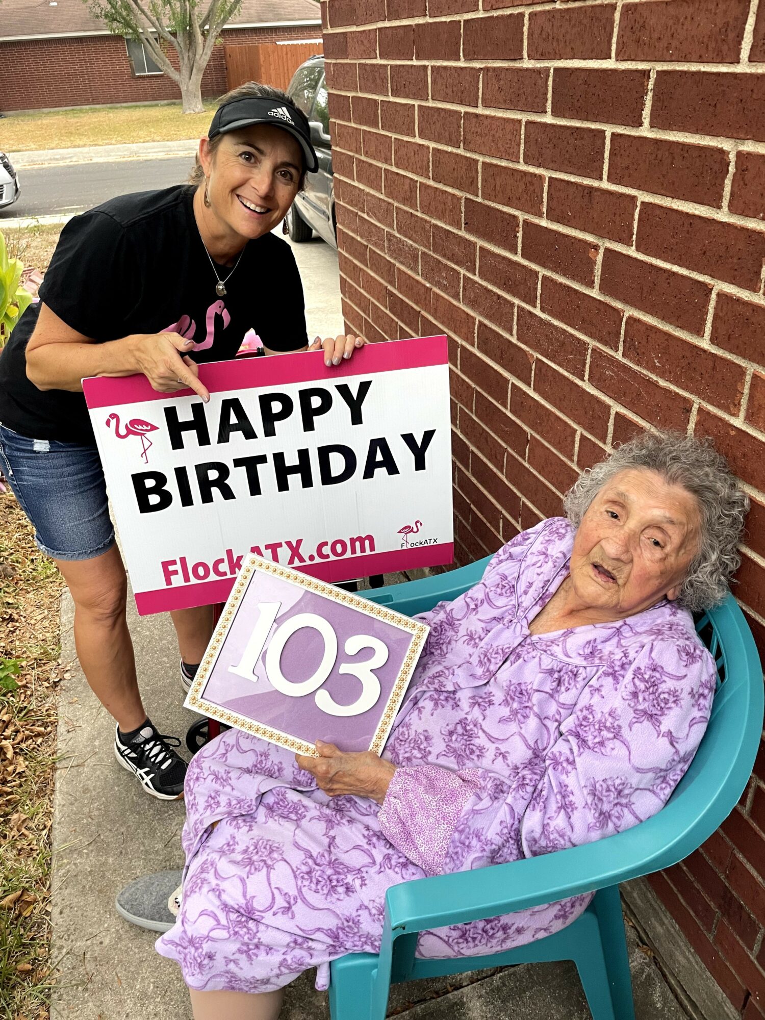 Young woman holding a birthday sign with an elderly woman in a chair holding a calendar, outside near a brick wall.