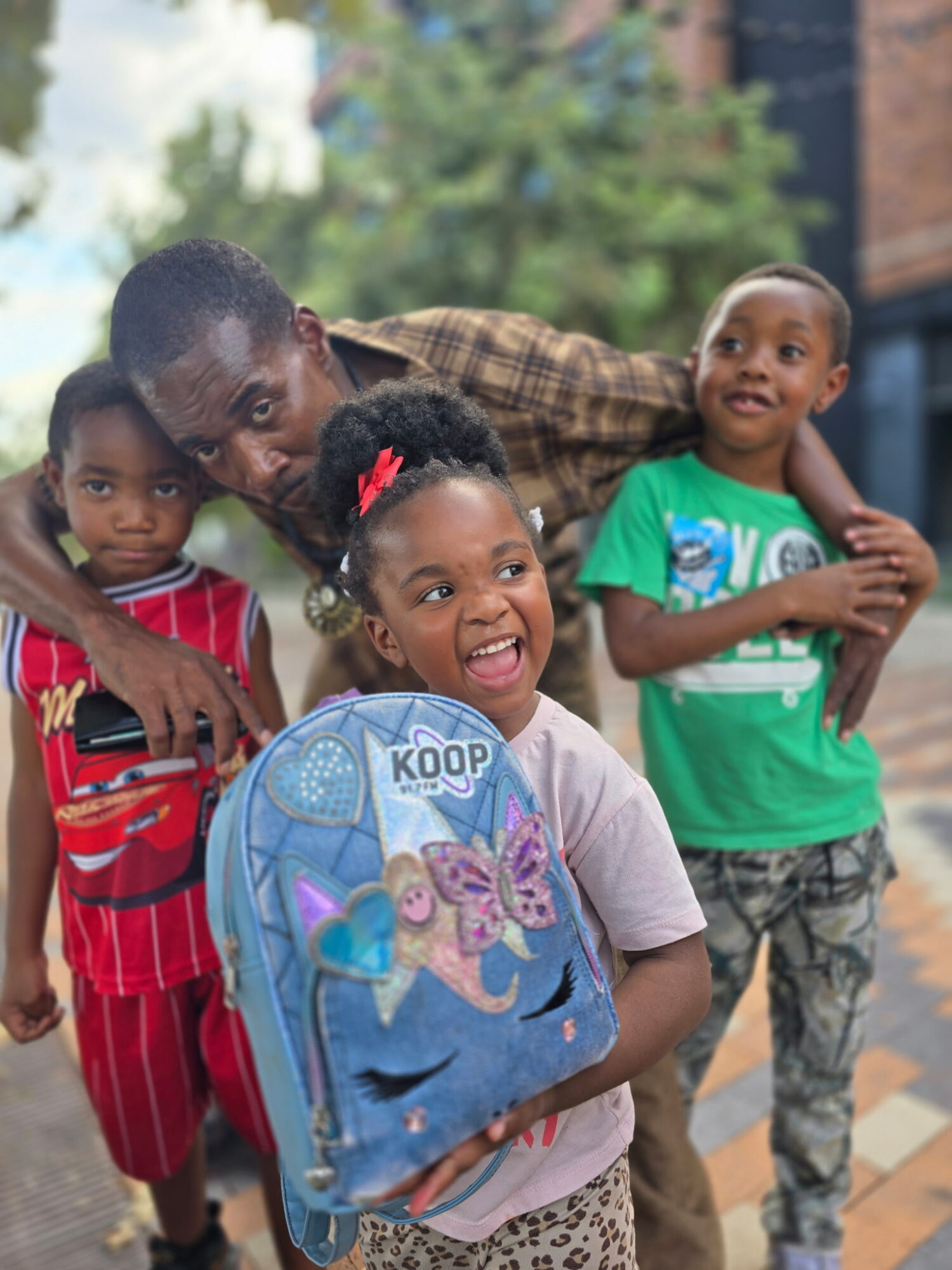 Four children and an adult outdoors, smiling and posing for the camera, with trees and buildings in the background.