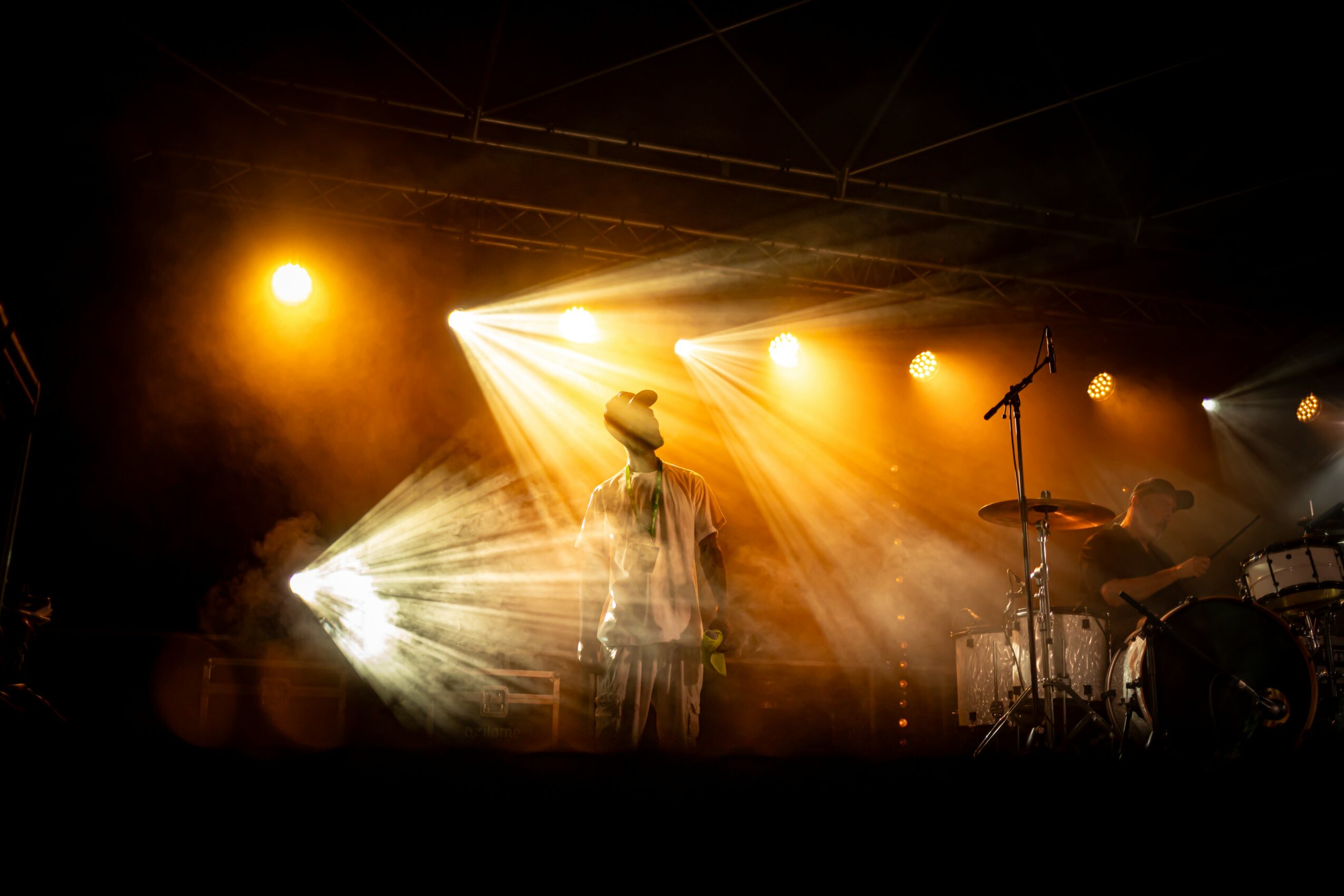 Stage with bright lights shining on a person, musical instruments visible, dark background, concert atmosphere.