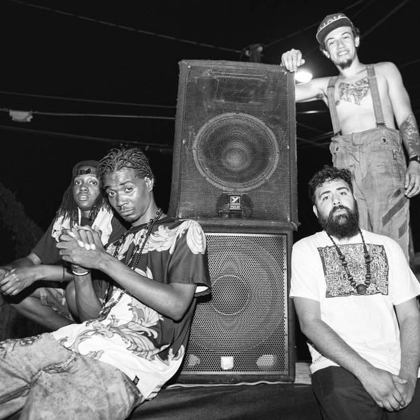 Four people around a large speaker, one standing on a platform, two sitting, one leaning on the speaker, at night.
