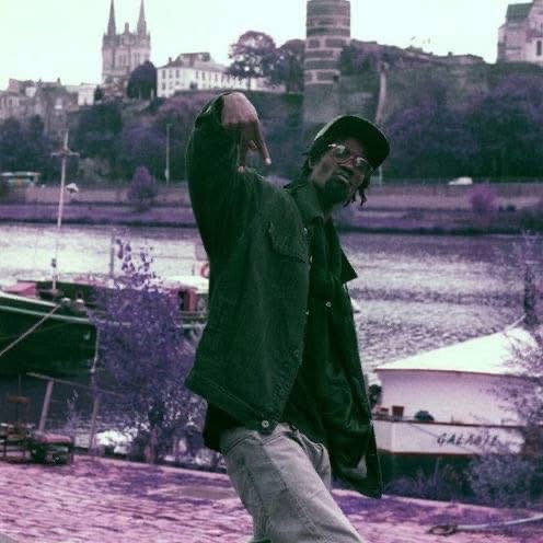 Person standing near a river with boats, cityscape, and historic buildings in background, wearing a cap and casual clothing.