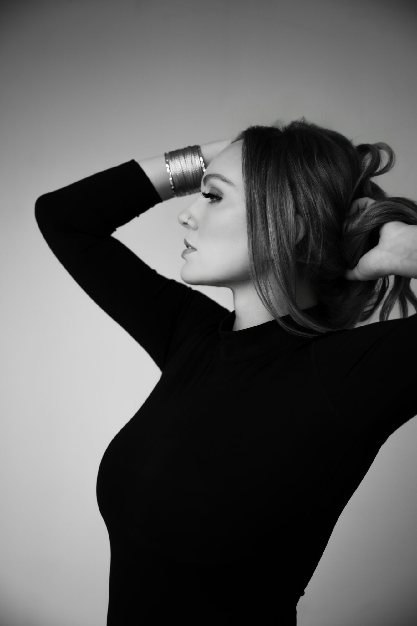 Woman with long hair, wearing a black top, poses with her hand on her head, looking to the side, in black and white.