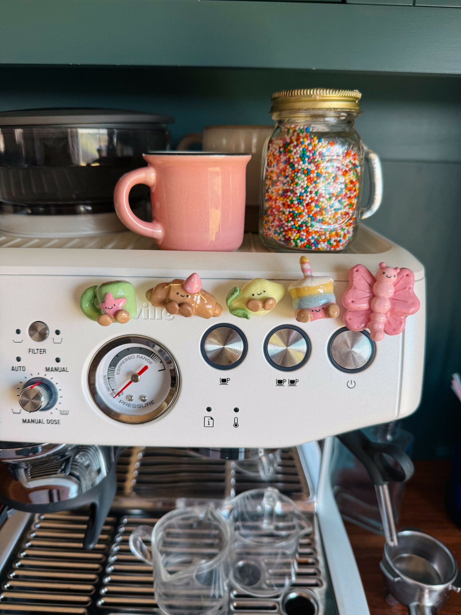 Coffee machine with pink mug, jar of colorful sprinkles, and small animal figurines on top, with cups and utensils below.