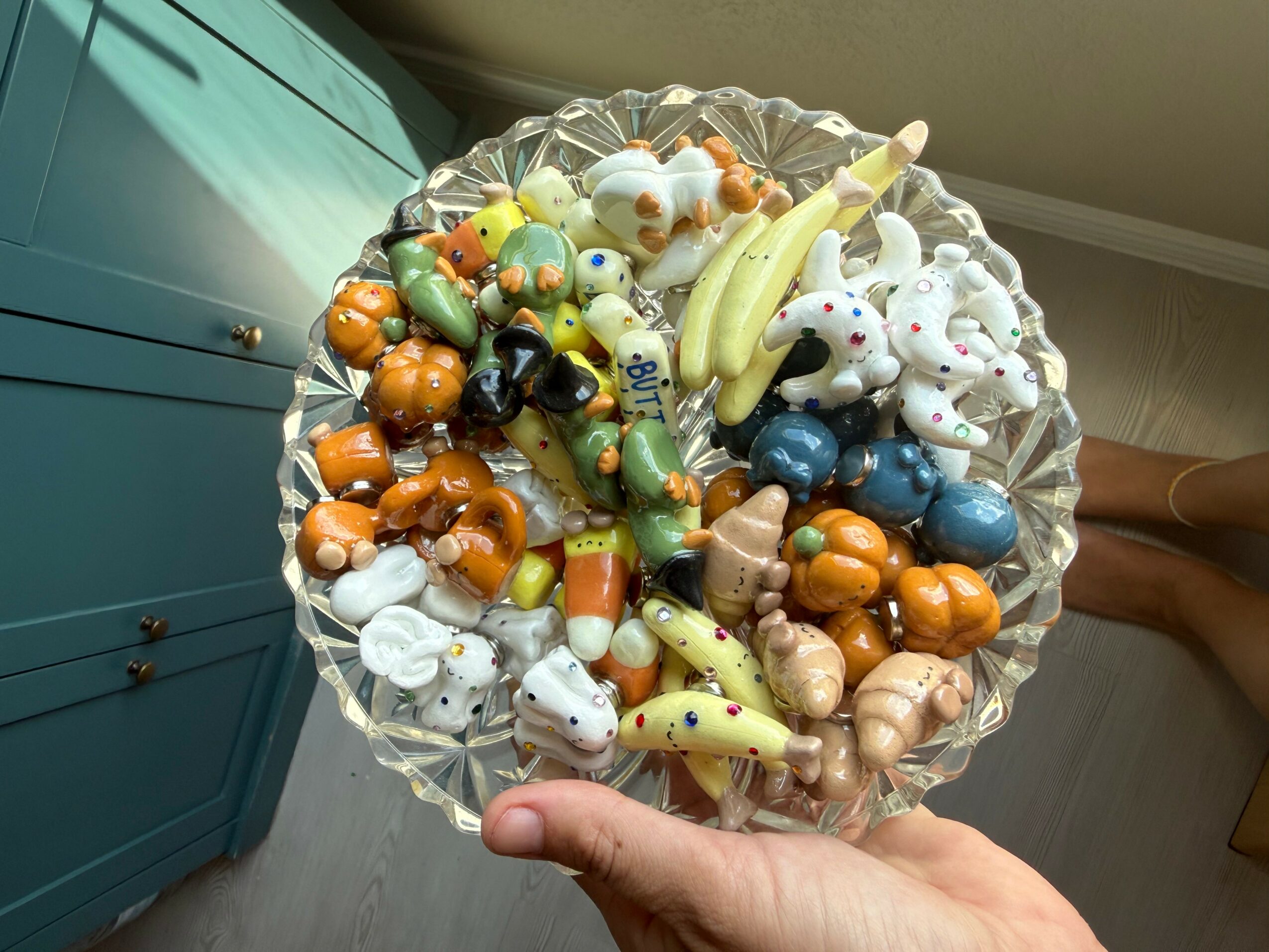 Hand holding a glass bowl filled with colorful candies and chocolates, with a blue cabinet in the background.