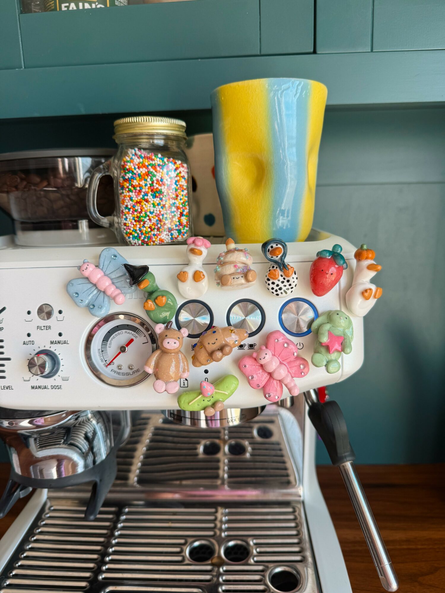 A coffee machine with colorful hair ties and a jar of sprinkles on top, and a yellow and blue mug behind.