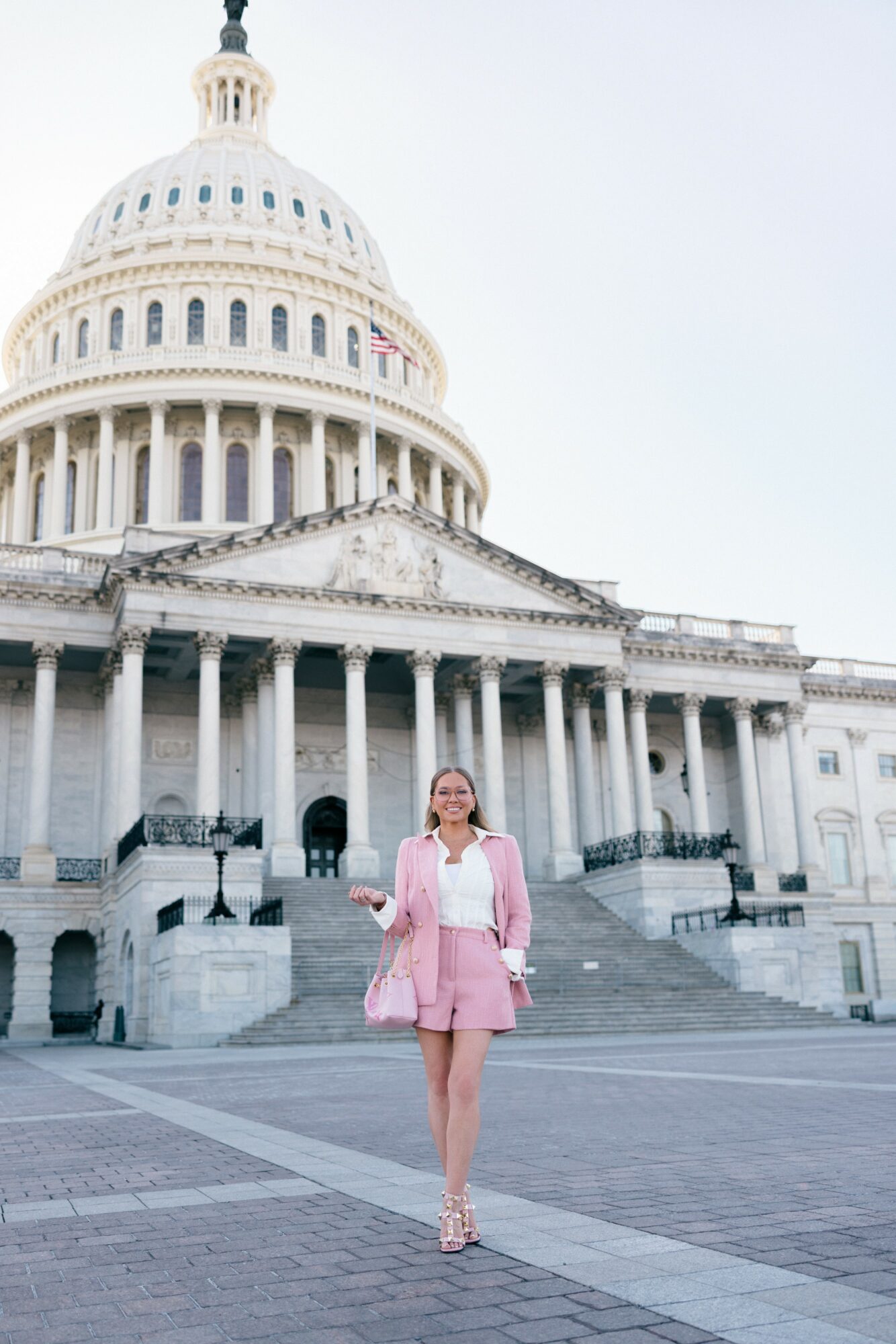 Woman in pink dress and jacket standing in front of the U.S. Capitol building, clear sky, steps behind her.