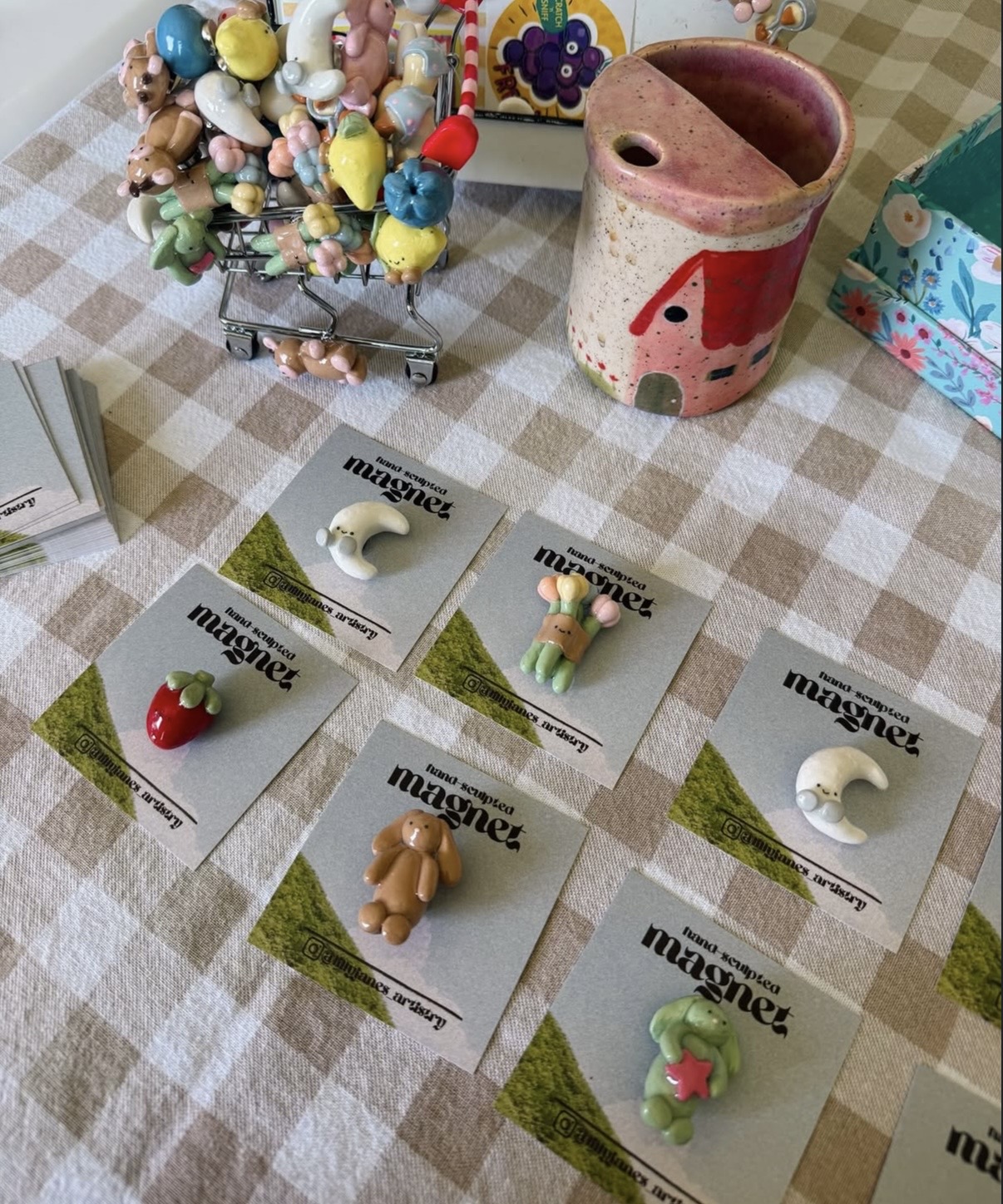 Six small figurines on cards arranged on a checkered tablecloth, with a pink ceramic container and a decorated box nearby.