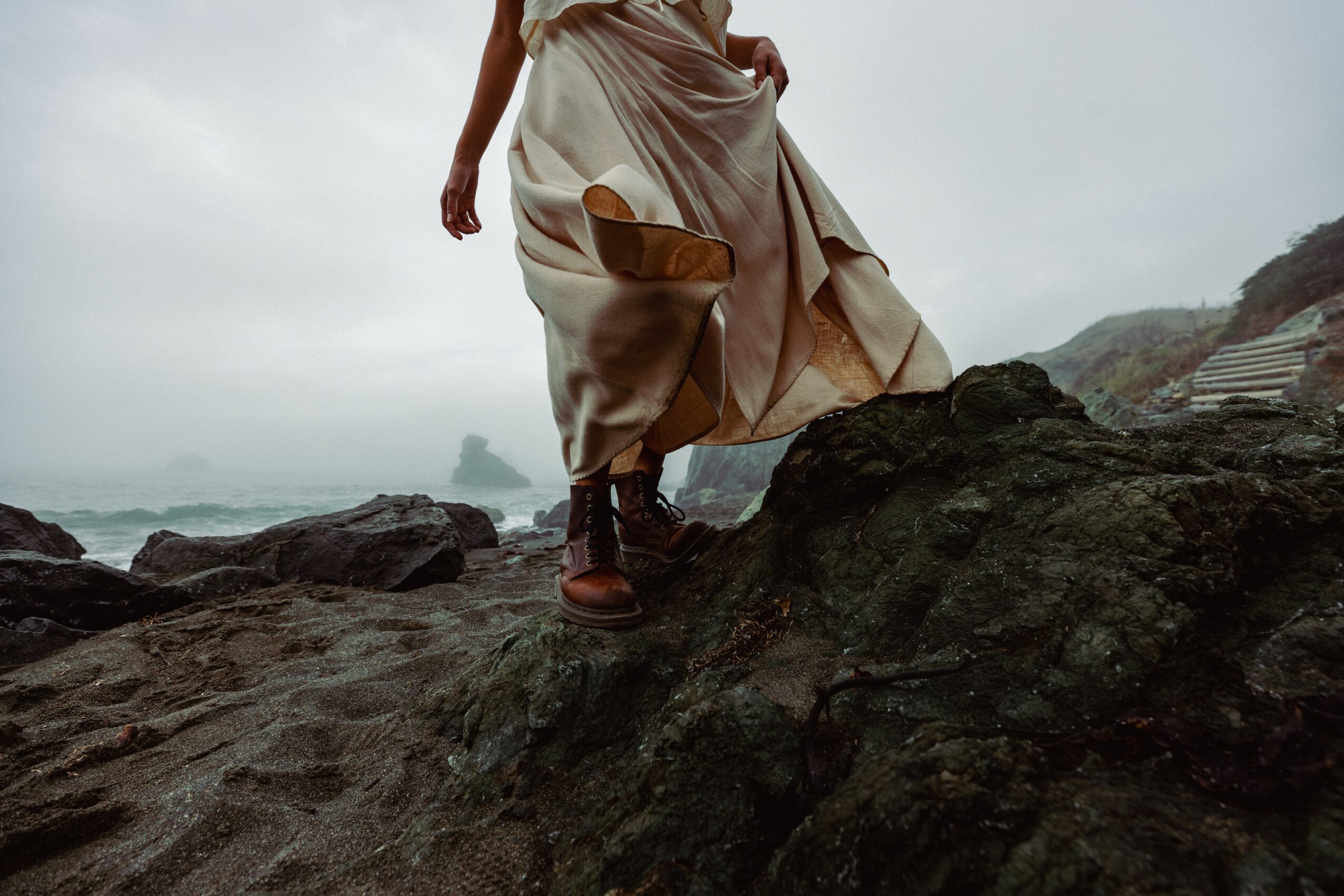 Person walking on rocks near the ocean, wearing a long dress and boots, with cloudy sky in background.