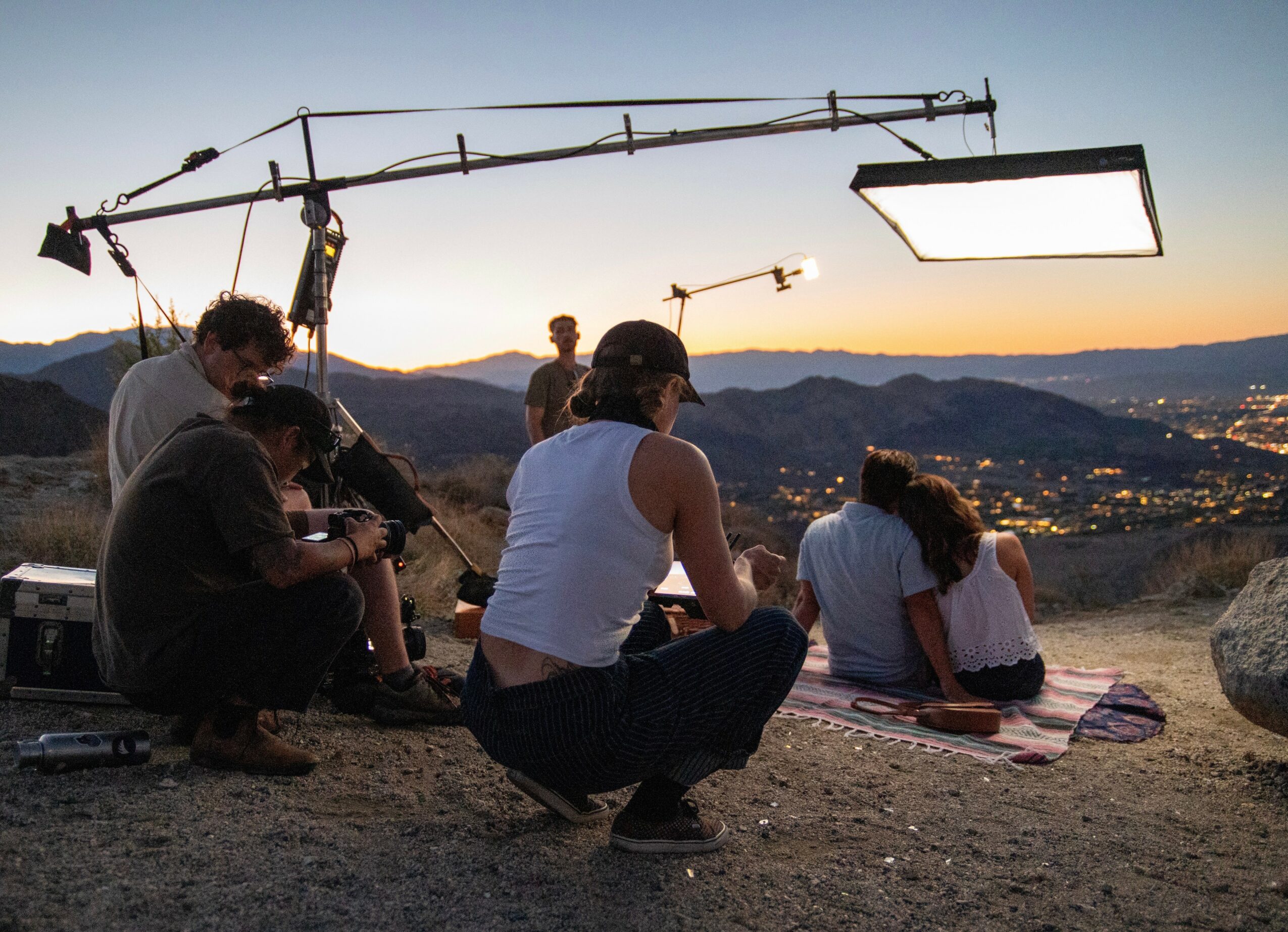 Filming crew with camera and lighting equipment outdoors during sunset, with people sitting on a blanket overlooking a valley.