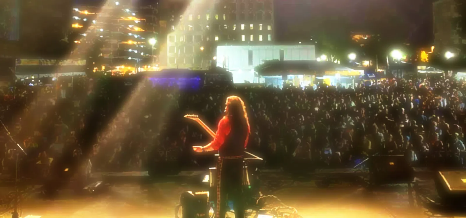 Performer on stage with a large crowd in the background, stage lights shining, nighttime setting.