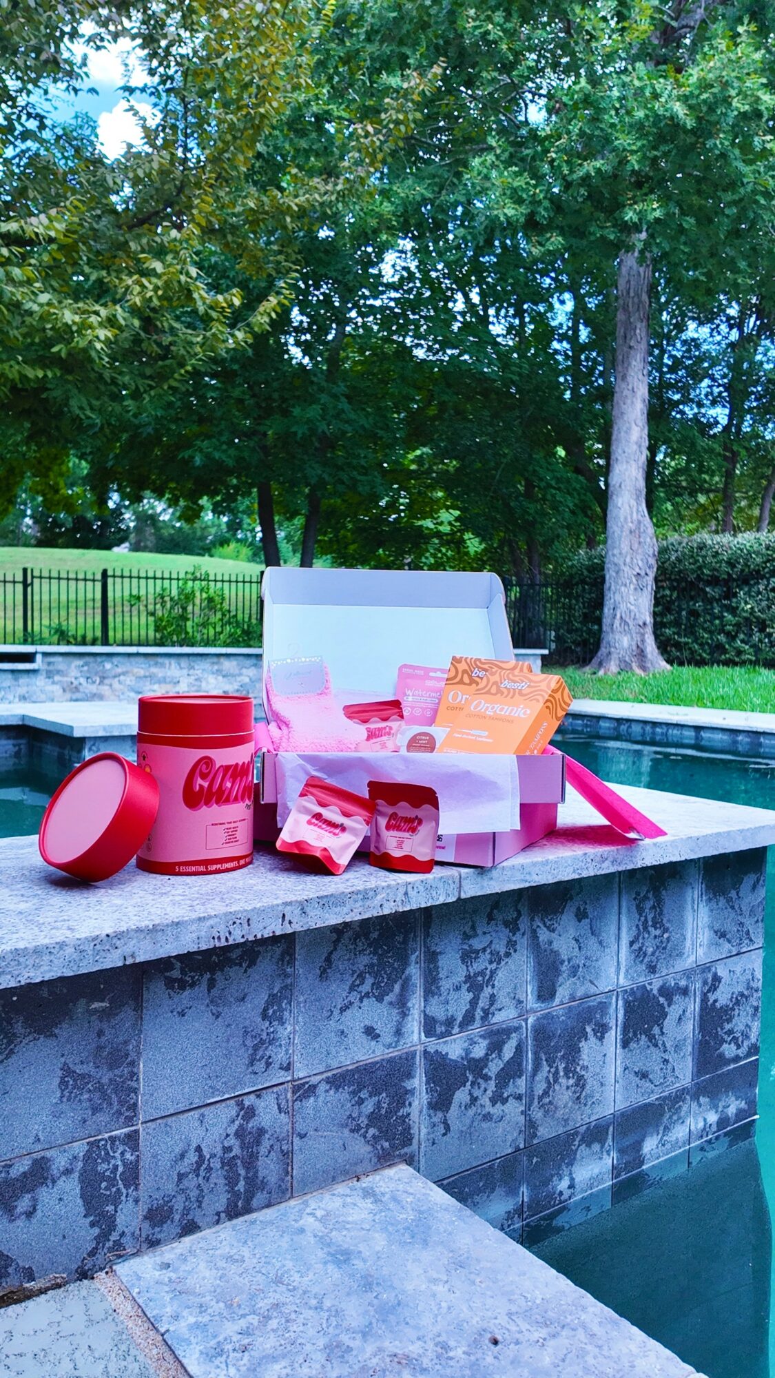 Poolside table with pink and red food items and a pink container, surrounded by trees and a fence in the background.