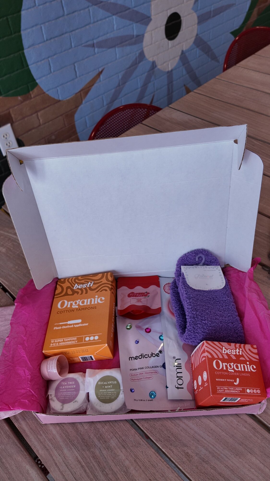 Open box containing organic soap, lotion, a purple washcloth, and other personal care items on a wooden surface.