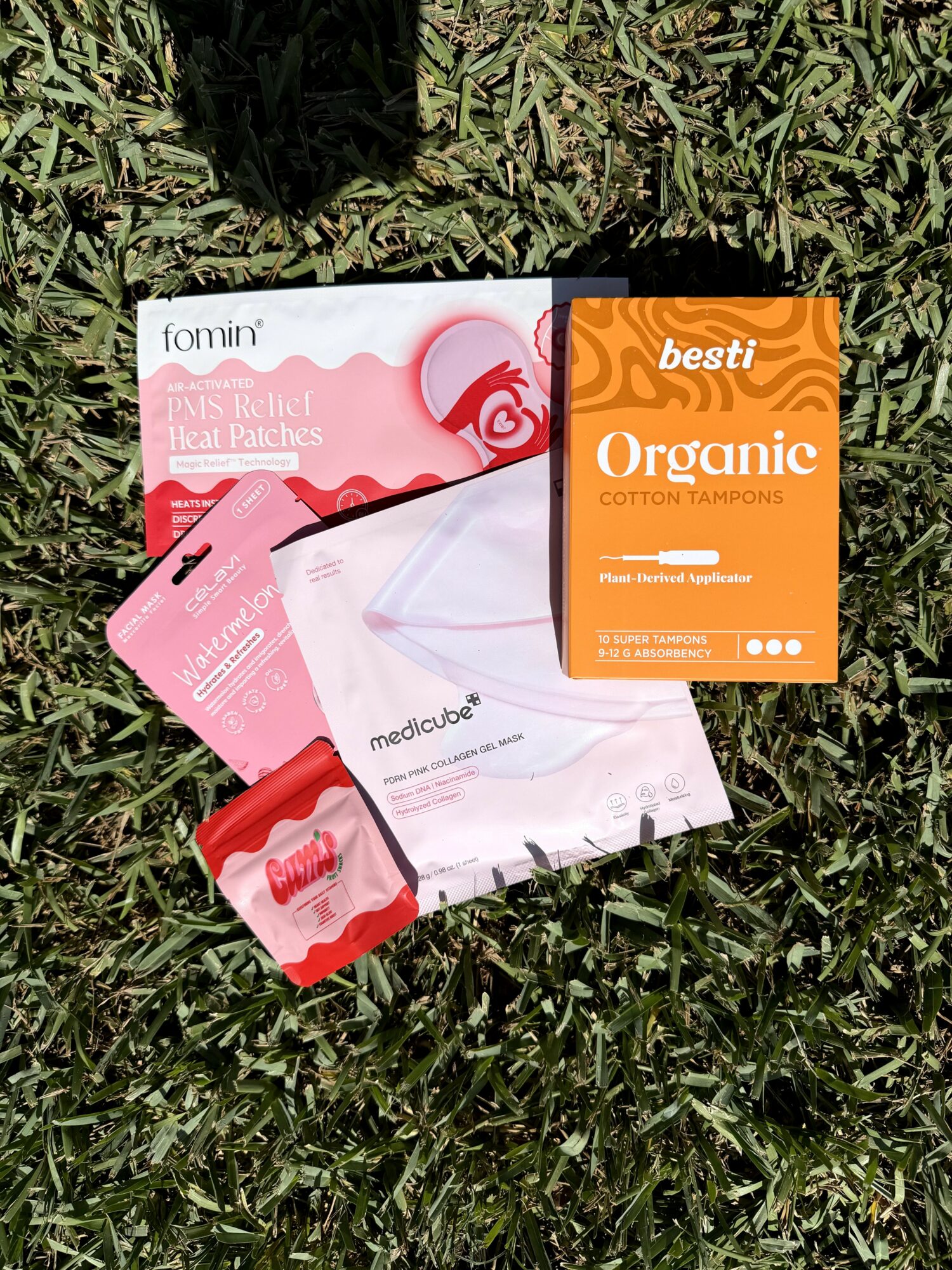 Various feminine hygiene products and packaging on grass, including pads and tampons, with orange and pink labels.