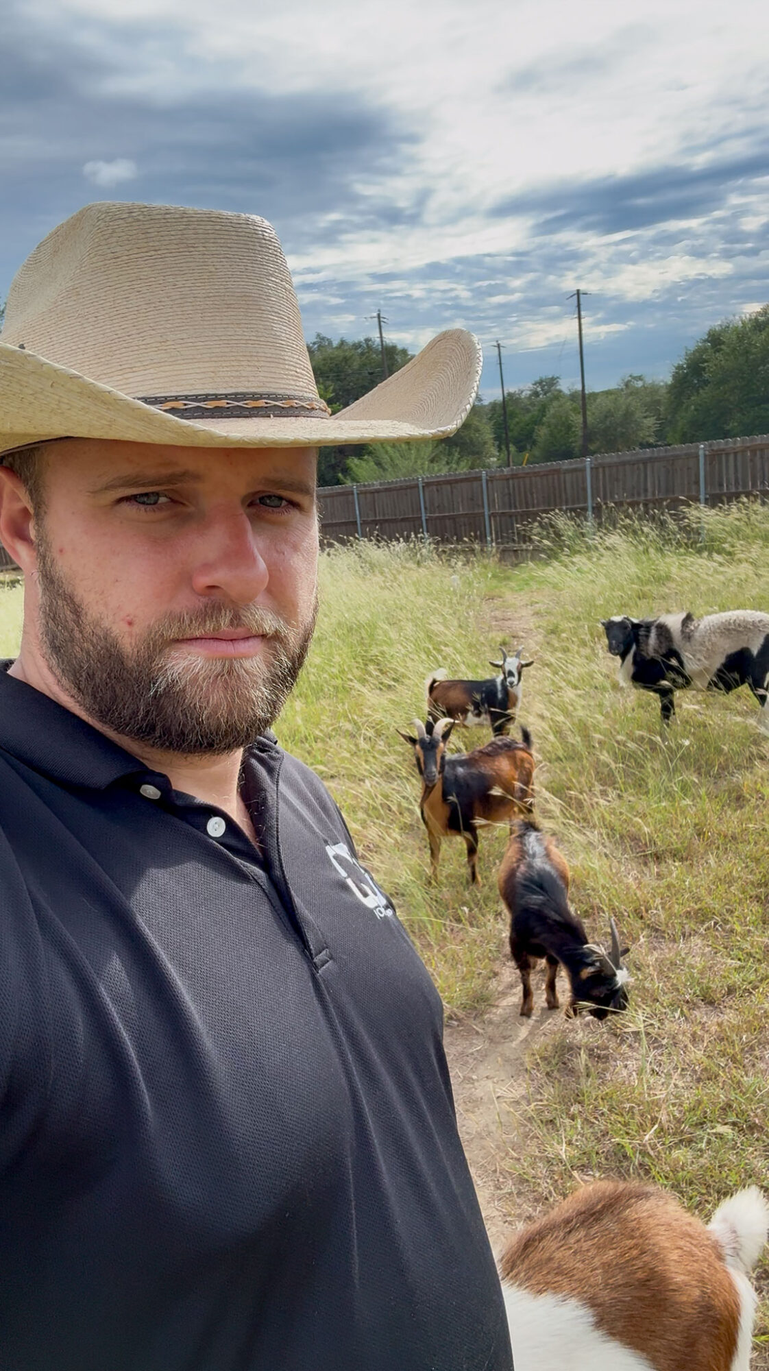 Man wearing a cowboy hat and dark shirt standing outdoors with cows in the background.
