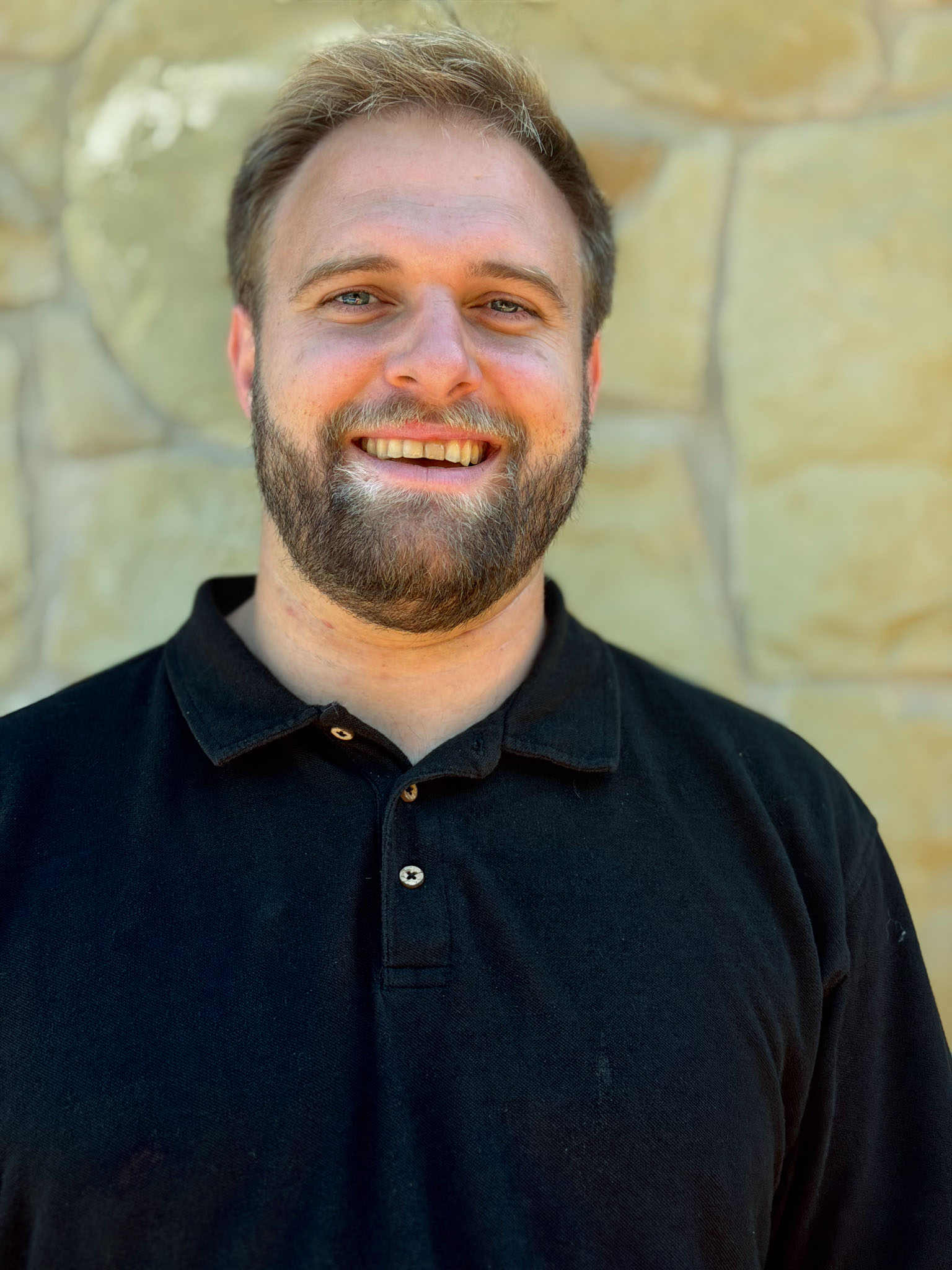 Smiling man with beard and short hair, wearing a black polo shirt, standing outdoors against a stone wall.