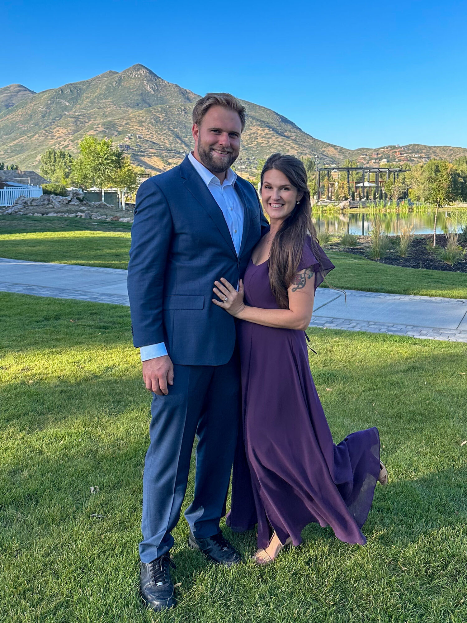 A man in a blue suit and a woman in a purple dress stand together outdoors with mountains and a lake in the background.