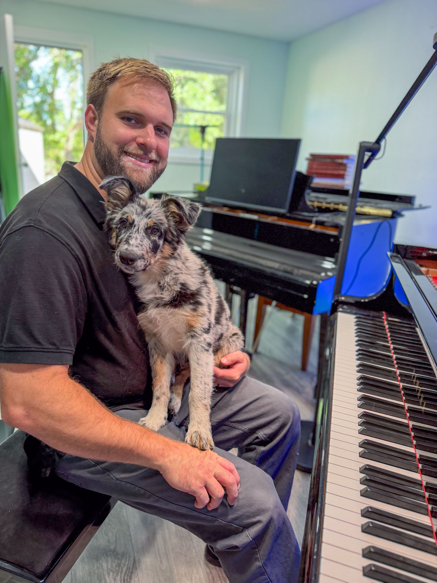 Man sitting at a piano holding a small dog, with a window and a laptop in the background.