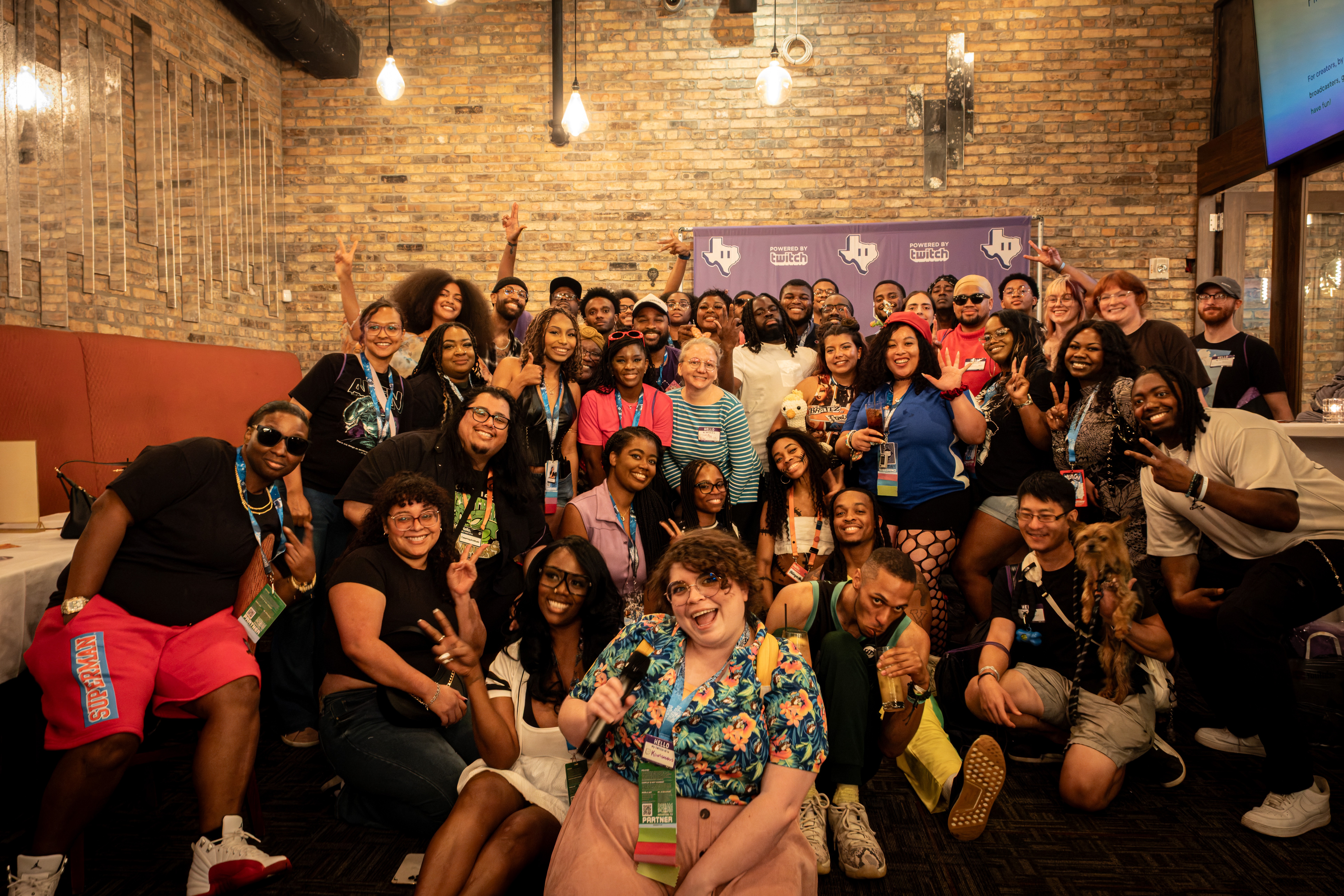 Group of diverse people gathered in a brick-walled venue, smiling and posing for a photo, some making peace signs.