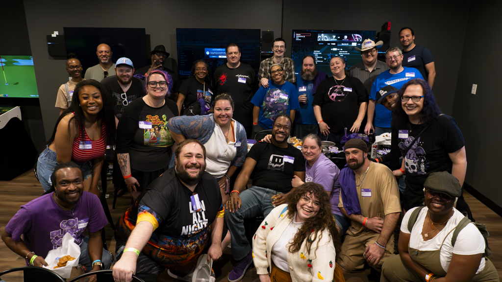 Group of people smiling and posing for a photo indoors, some wearing glasses and casual clothing.