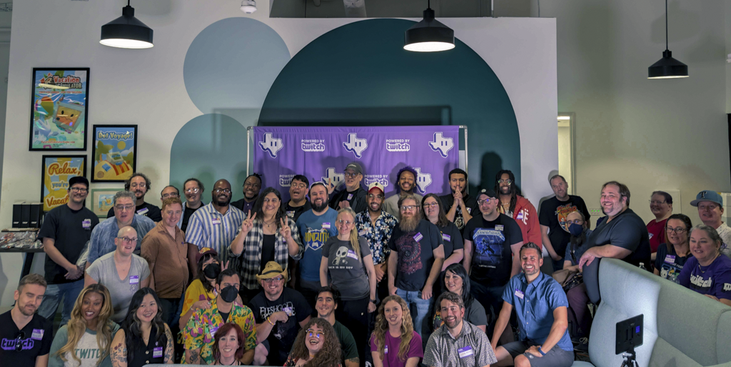 Group of people gathered indoors in front of a purple backdrop with logos, some standing and some sitting, smiling.