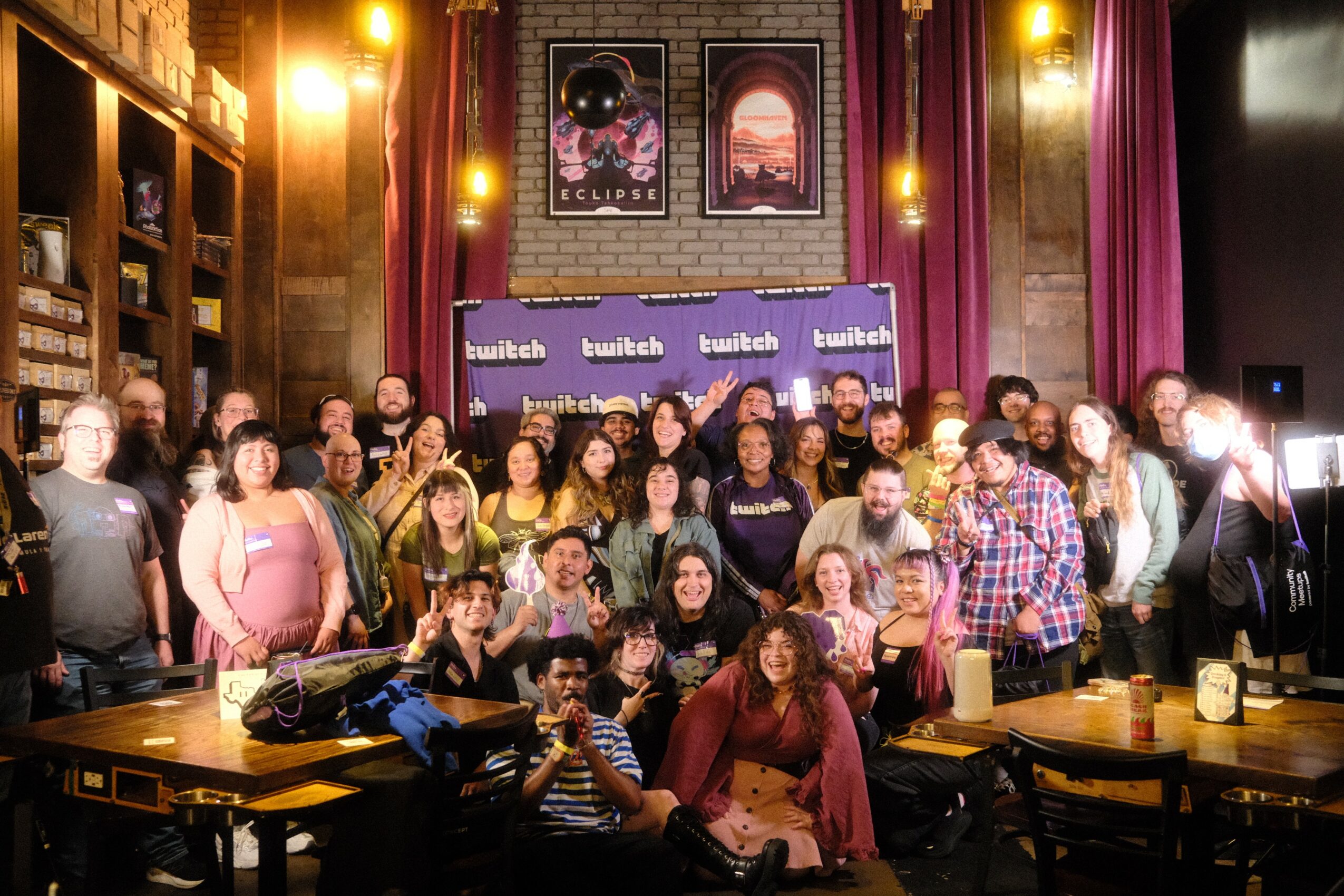 Group of people gathered in a room with bookshelves, posters, and a banner, some sitting and some standing, with tables and drinks.