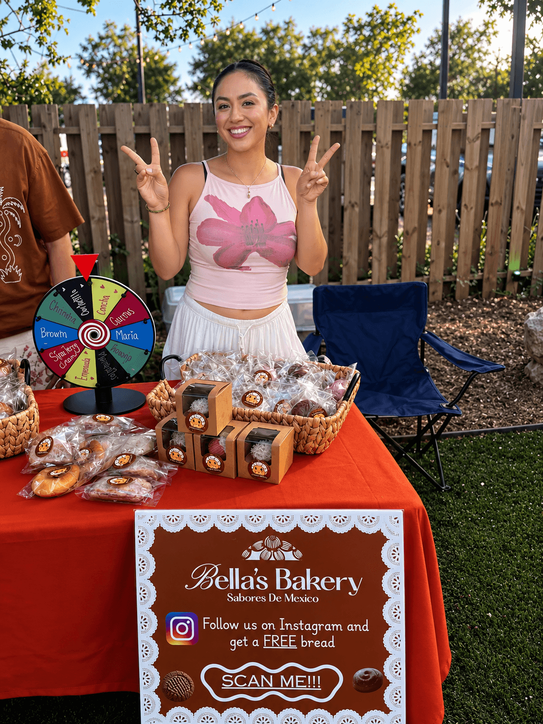 Smiling woman at bakery booth making peace signs, table with baked goods, sign with bakery info, outdoor setting.