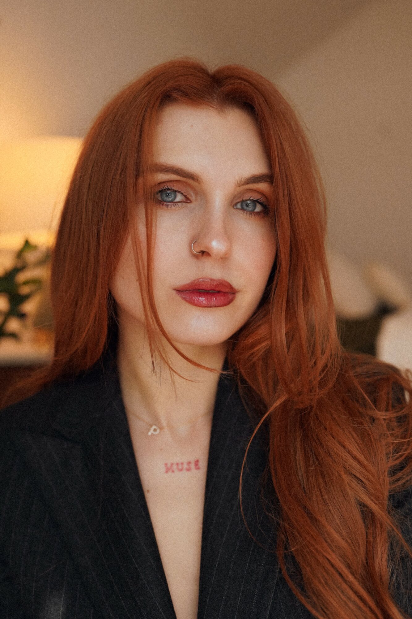 Close-up of a woman with long red hair, blue eyes, and red lipstick, wearing a dark top, indoors.