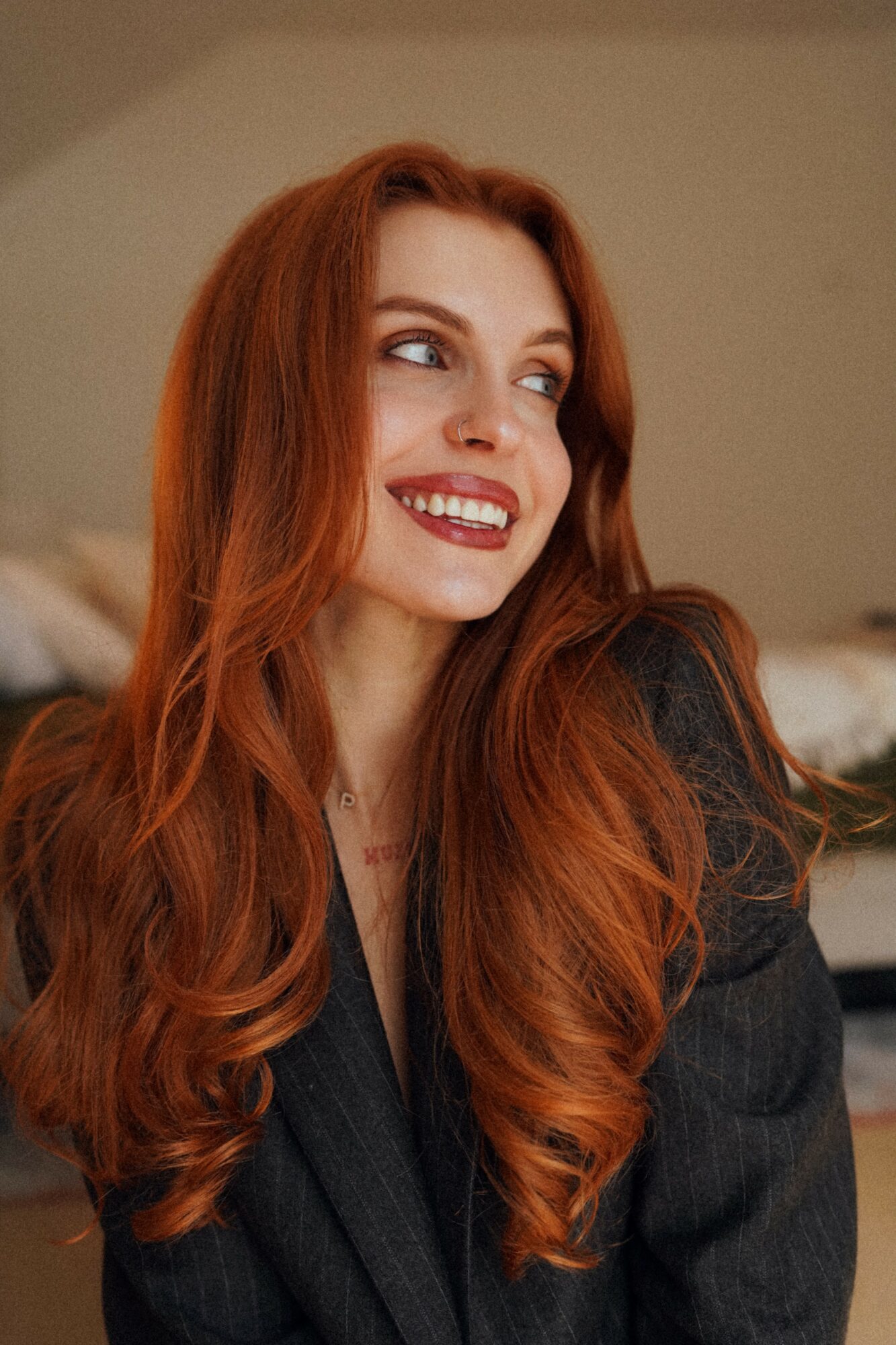 Woman with long red hair smiling, looking to the side, wearing a dark blazer, indoors.