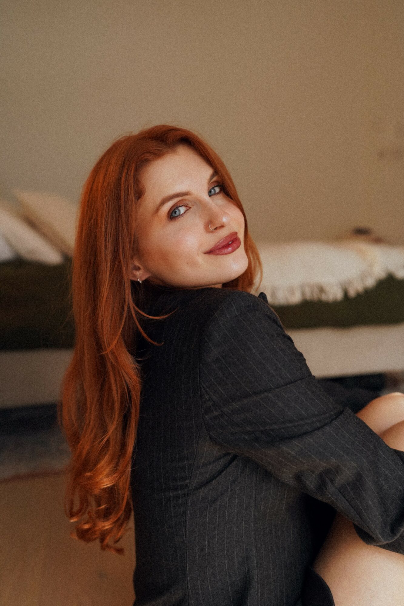 Woman with long red hair and makeup, wearing a dark top, sitting indoors with a bed in the background.