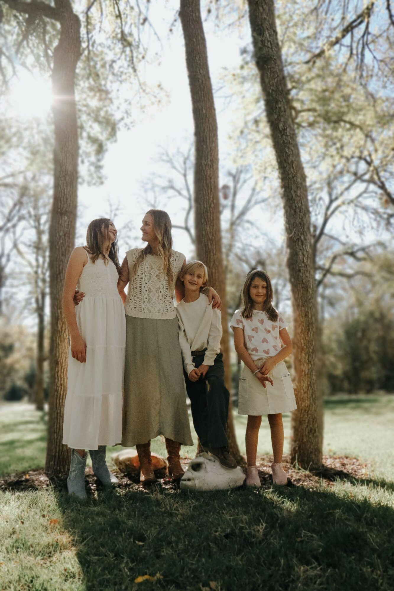 Four girls standing outdoors among trees, sunlight filtering through, dressed in light-colored clothing, smiling and looking at each other.
