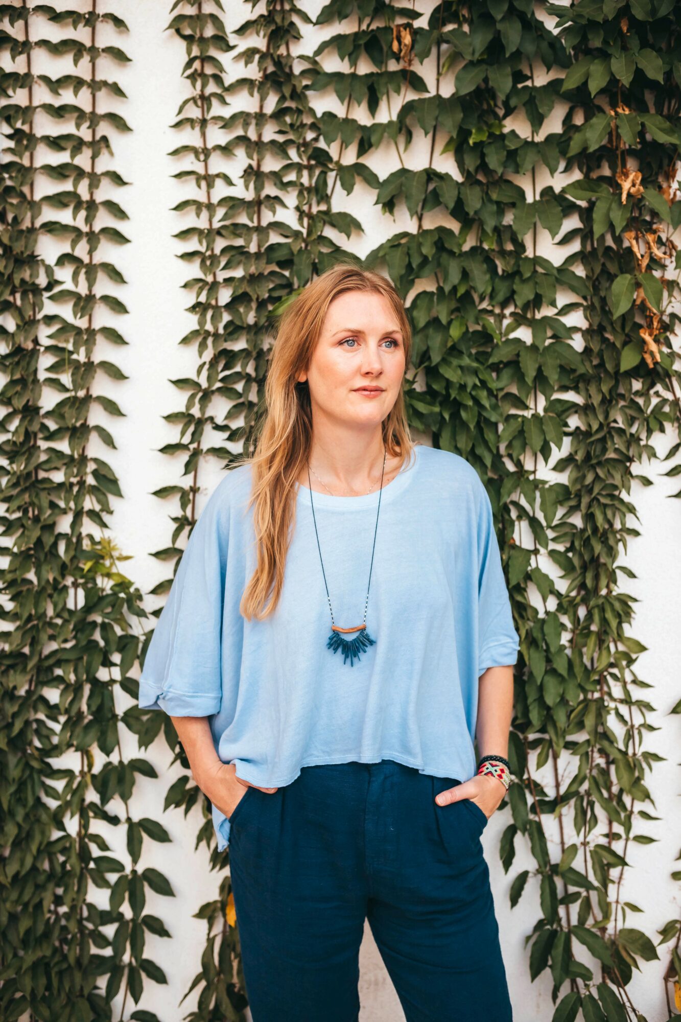 Woman with long hair wearing a light blue top and dark pants stands in front of green leafy vines.