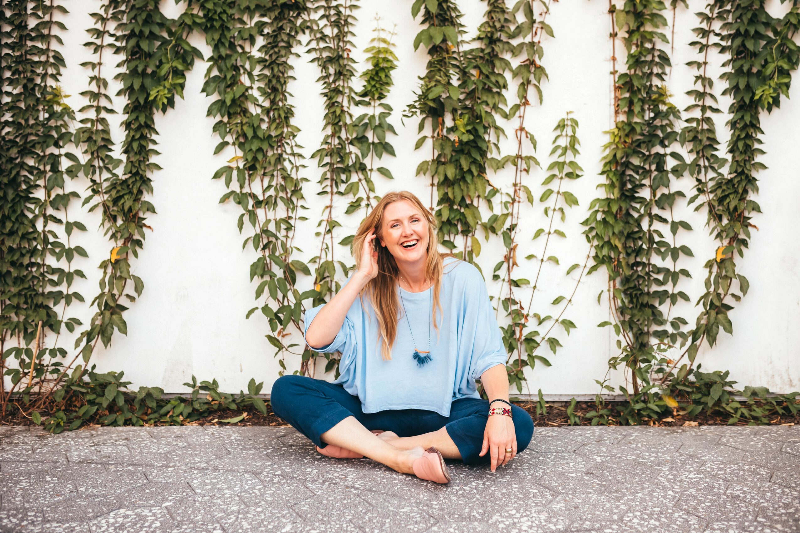 Woman sitting on ground in front of tall green plants, smiling and talking on phone, wearing a light blue top and dark pants.