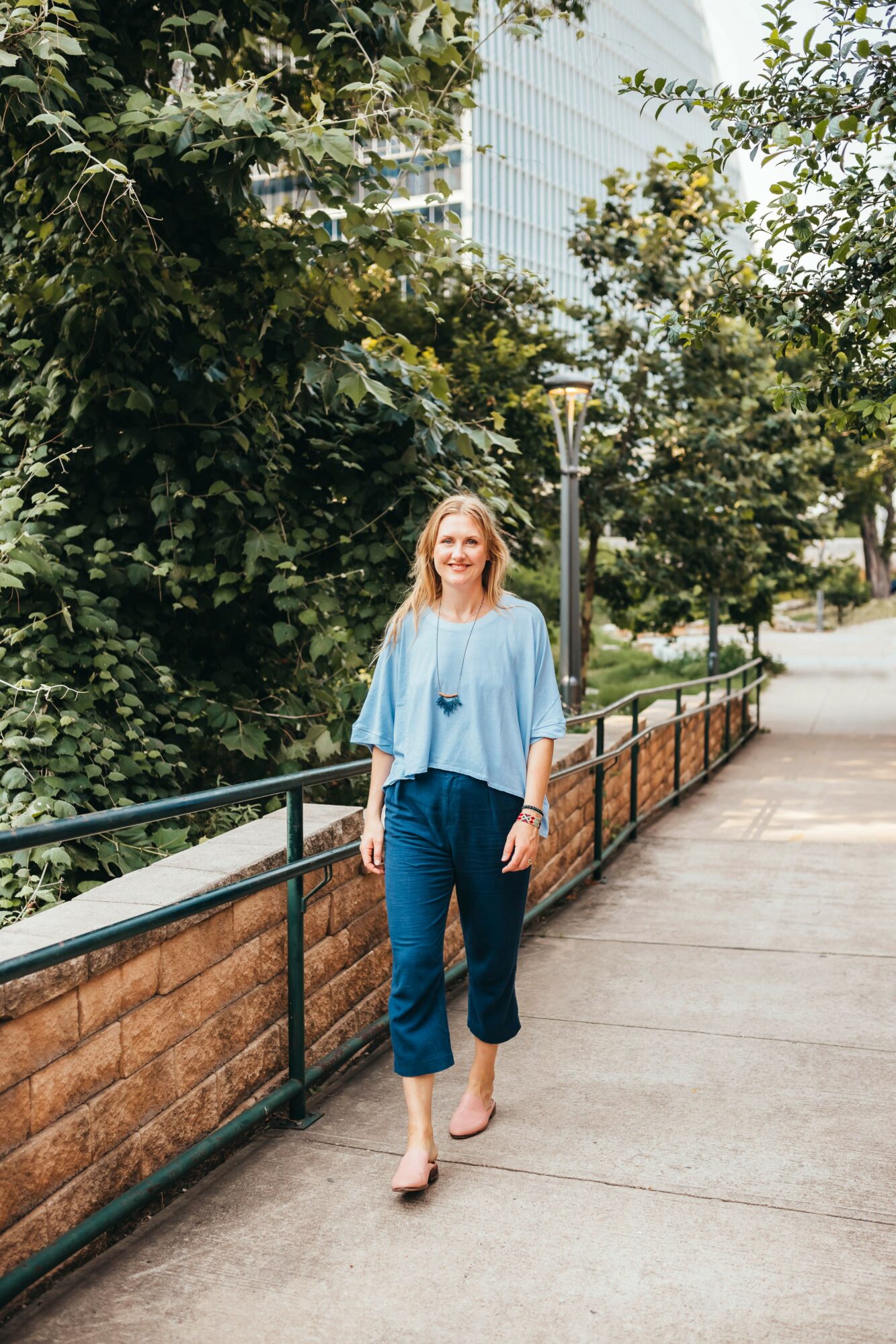 Woman walking on sidewalk with trees and buildings in background, smiling, wearing a light blue top and dark pants.