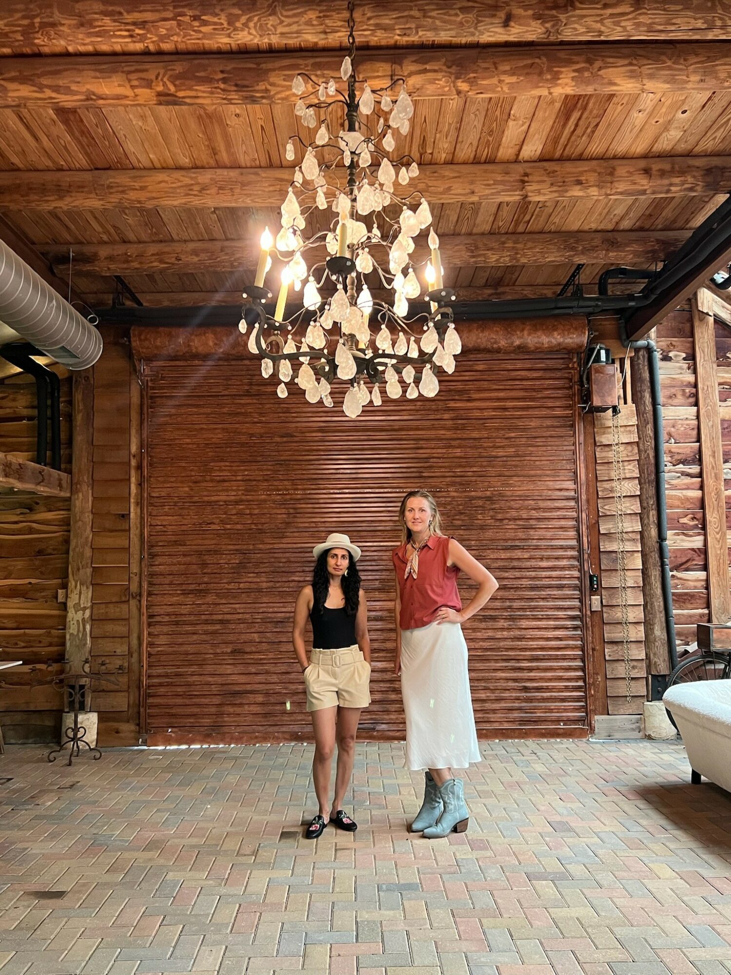 Two women stand under a chandelier in a rustic wooden room, one wearing a hat and shorts, the other in a long skirt and sleeveless top.