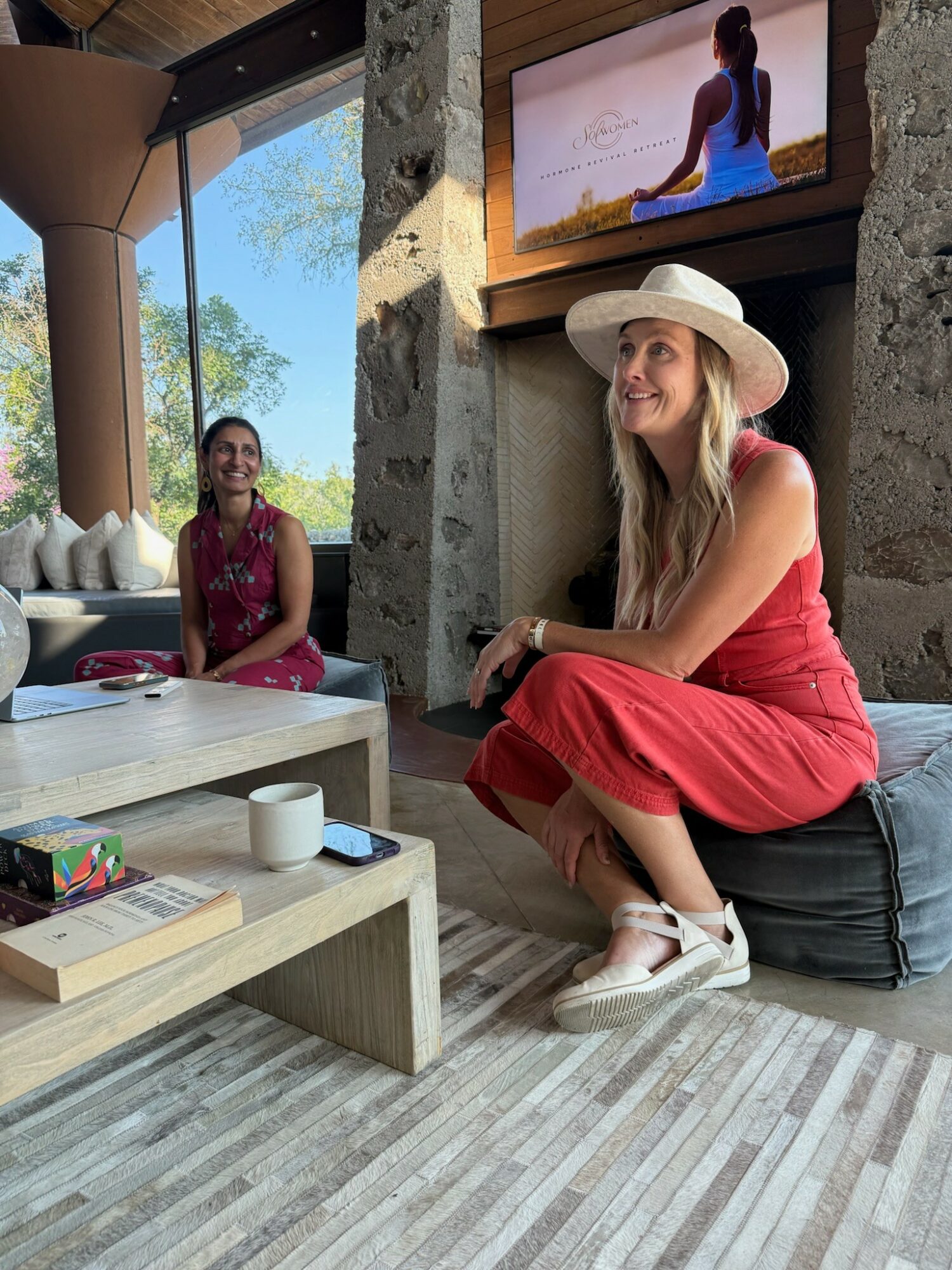 Two women sitting indoors, one on a cushion and the other on a table, with a TV and large window showing trees outside.