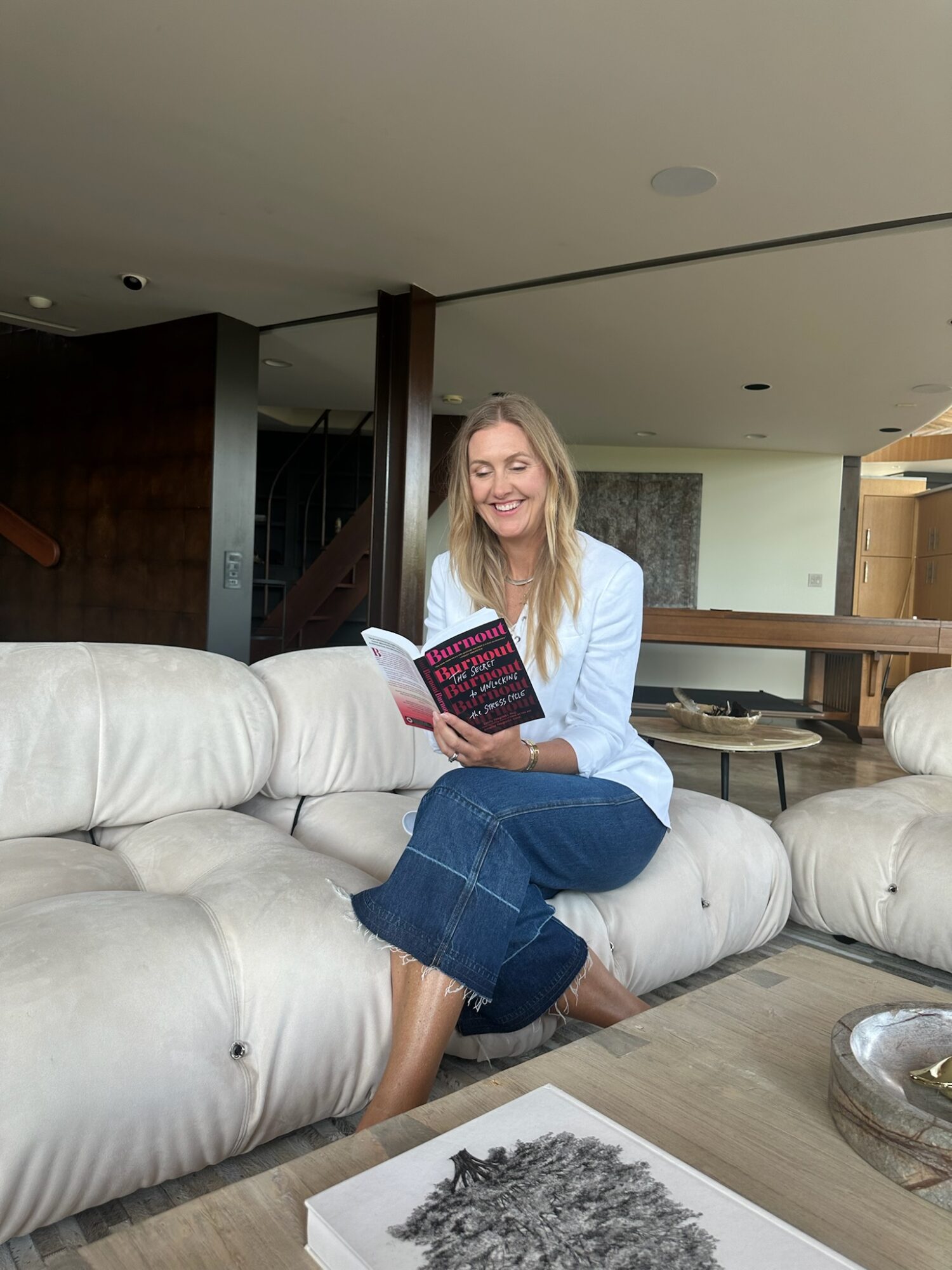 Woman sitting on a white sofa, reading a book, smiling, in a modern living room.