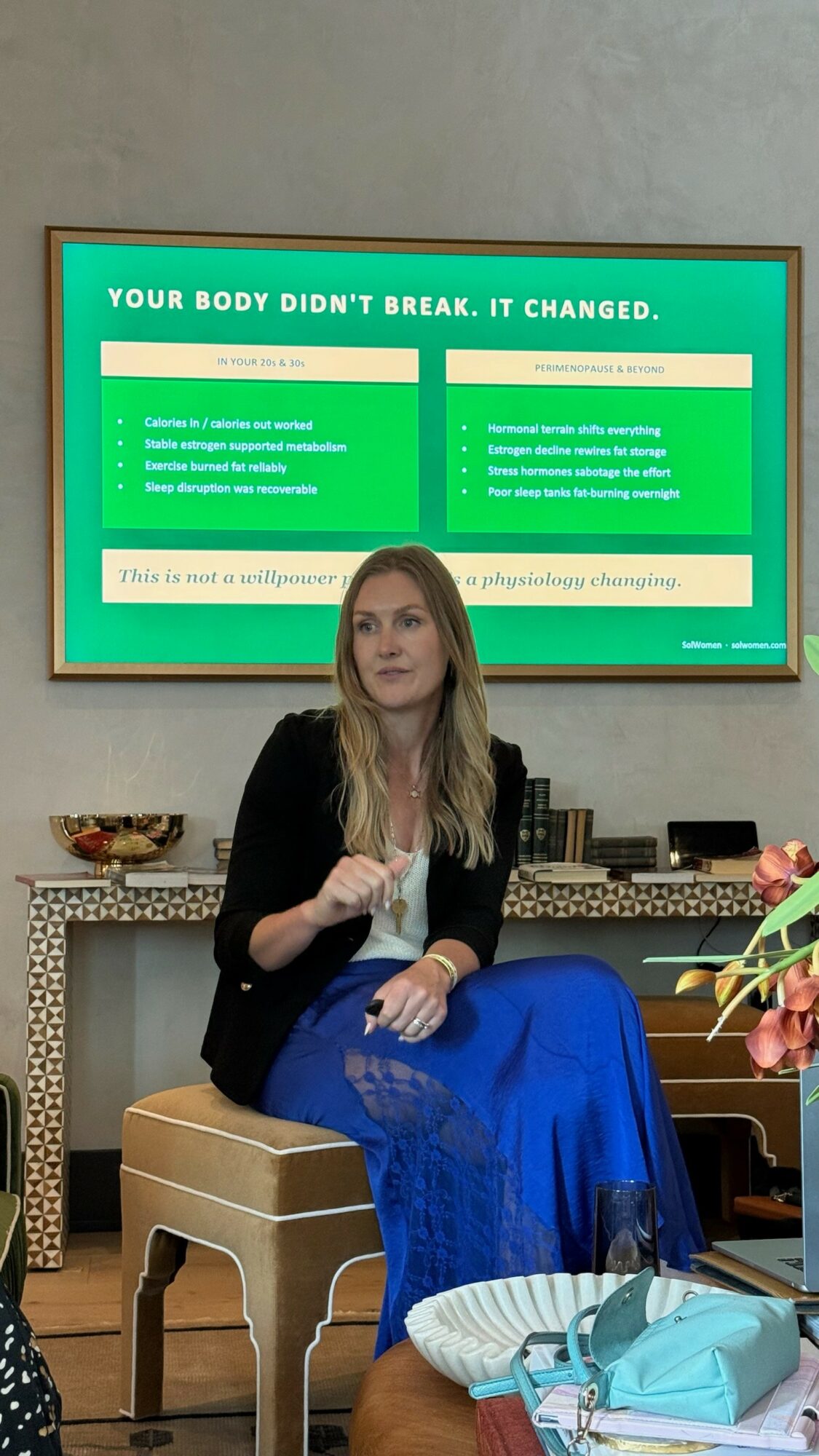 Woman sitting on a chair in front of a green screen with text, holding a pen, with a table and decorative items nearby.