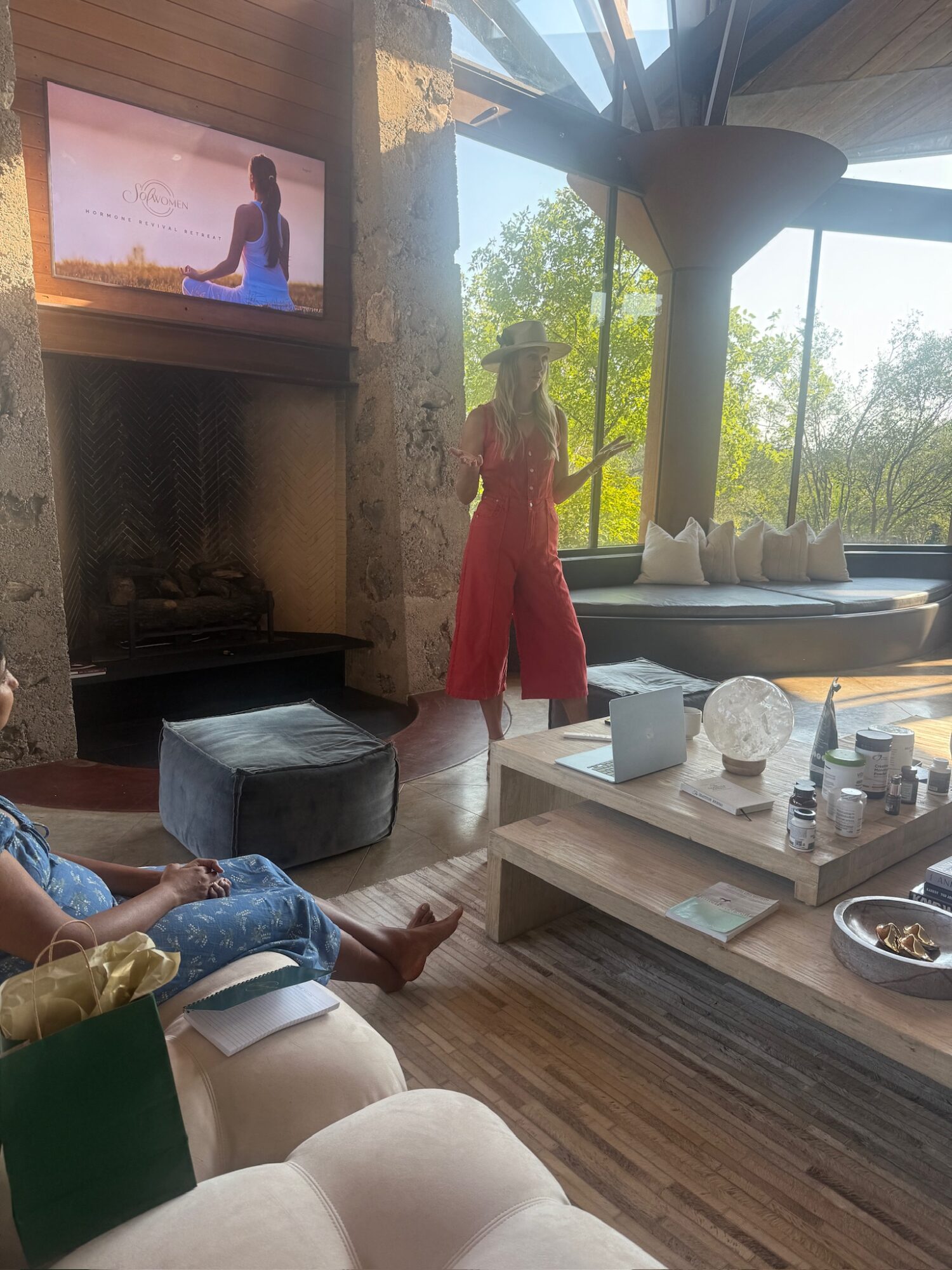 Living room with large windows, a woman in a red dress and hat, a person sitting on a sofa, and a TV screen showing a person outdoors.