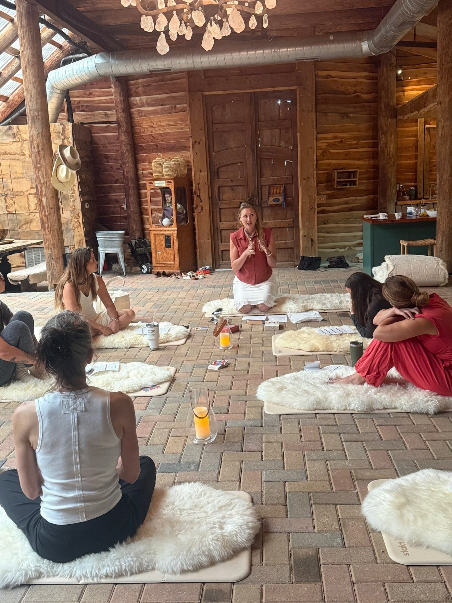 Group of people sitting on fluffy rugs in a rustic wooden room, with a woman leading a meditation or yoga session, drinks on the floor.