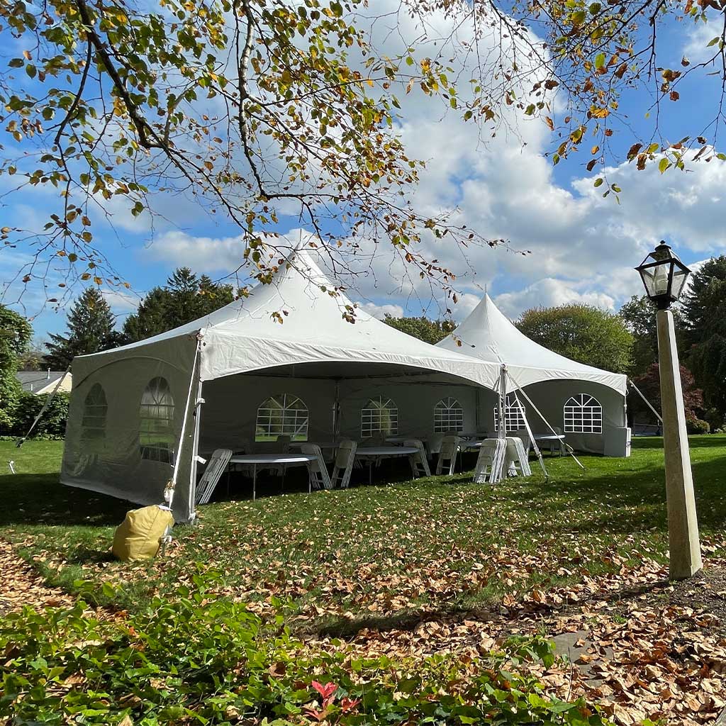 Large white event tent set up outdoors on grass with trees and a lamppost nearby, under a partly cloudy sky.