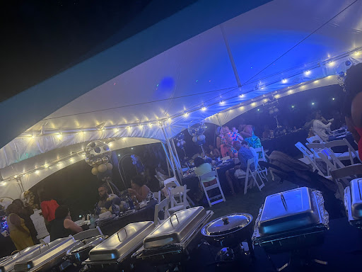 Nighttime outdoor event with tables, chairs, string lights, and people socializing under a canopy, illuminated by blue and warm lights.