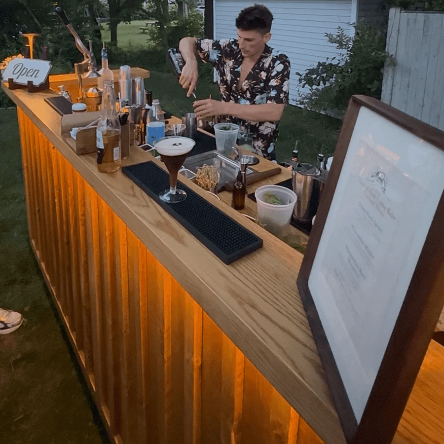 Person preparing drinks at outdoor bar with bottles, glasses, and a menu, illuminated by warm lighting, in a backyard setting.