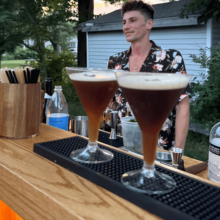 Person standing outdoors behind a bar with two cocktails in the foreground, trees and a building in the background.